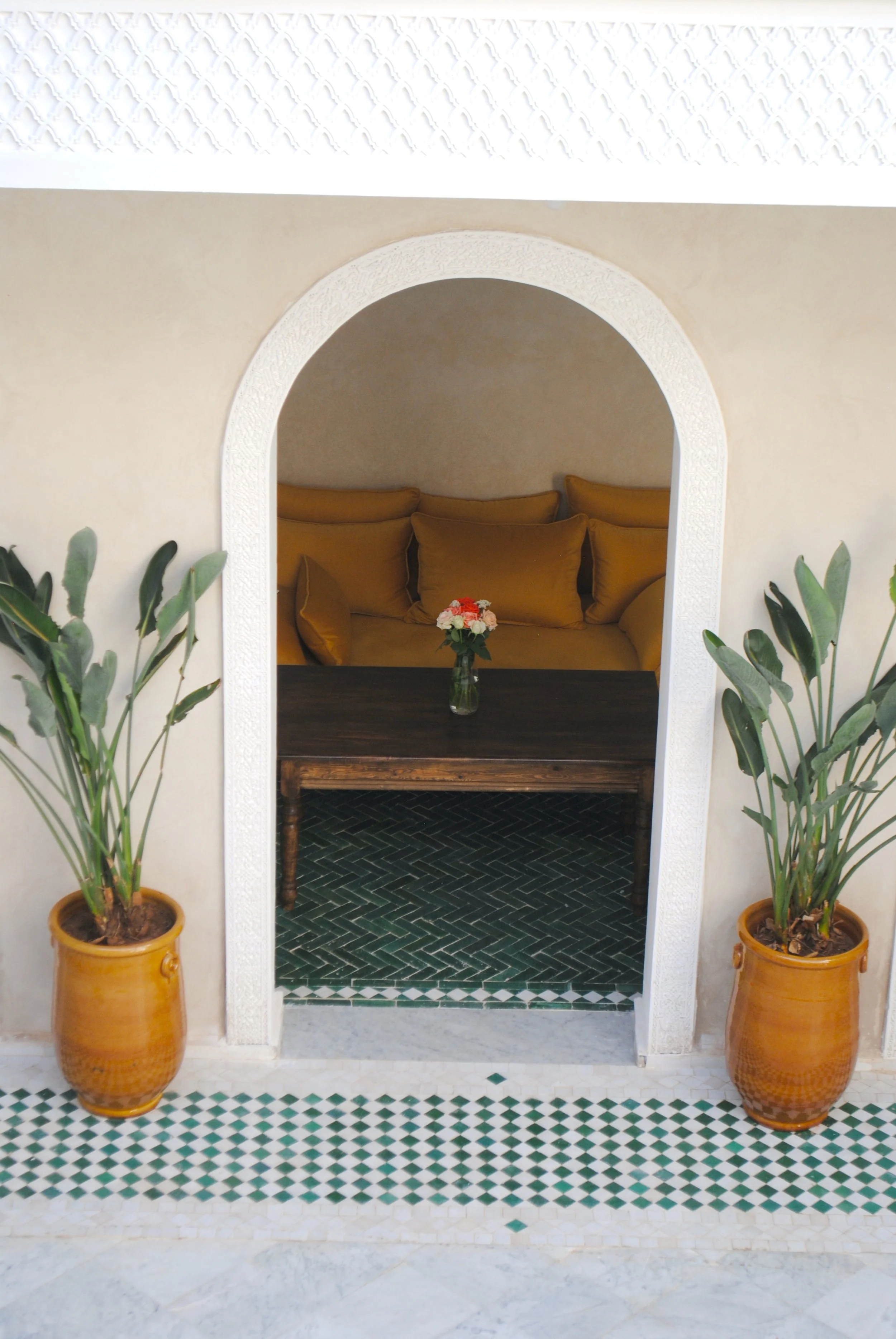 A decorative entryway with two potted plants on either side, leading to a cozy nook with a dark wood table, a small flower arrangement, and a yellow couch with several cushions.