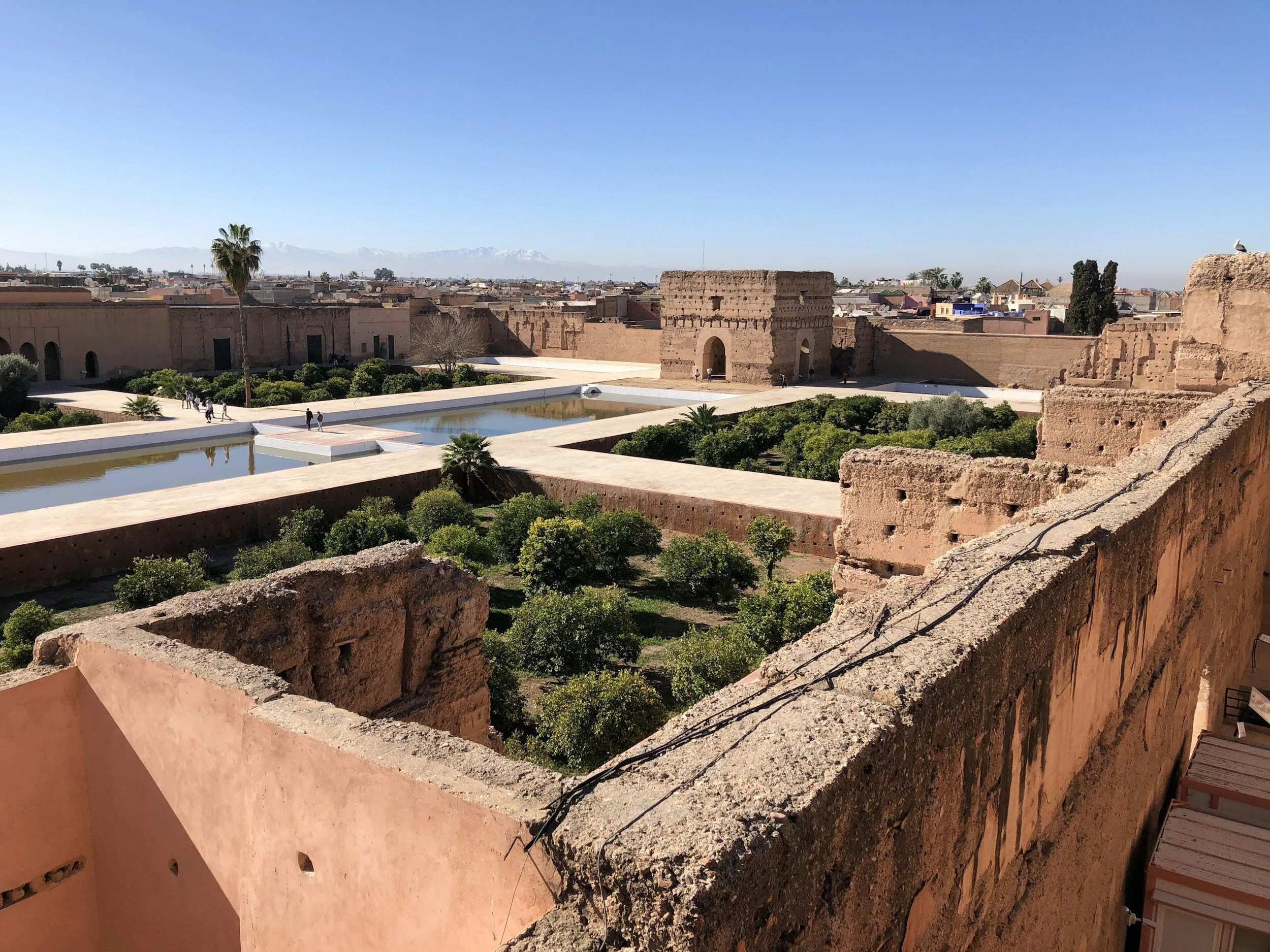 A historical Moroccan cityscape with ancient reddish-brown walls and tower, green garden with bushes and trees, and two rectangular pools of water in the courtyard under a clear blue sky.
