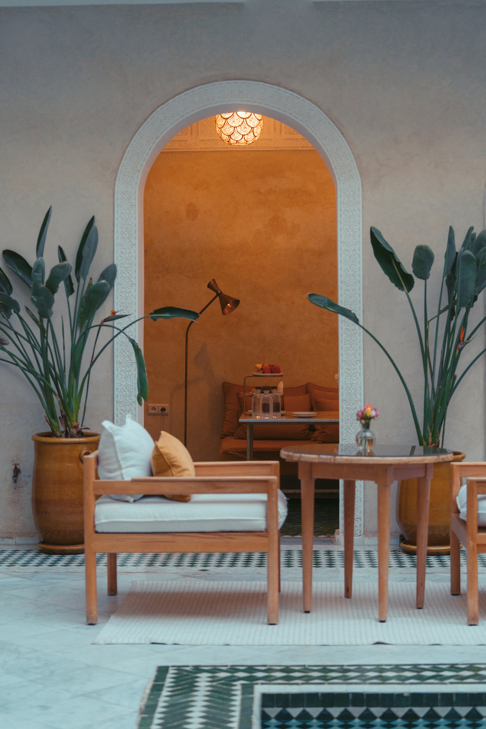 A narrow outdoor corridor with a tiled green and white patterned floor, wooden window frames with ornamental iron grilles, a hanging lantern, and a wooden door, with a building featuring a Moroccan or Andalusian architectural style.