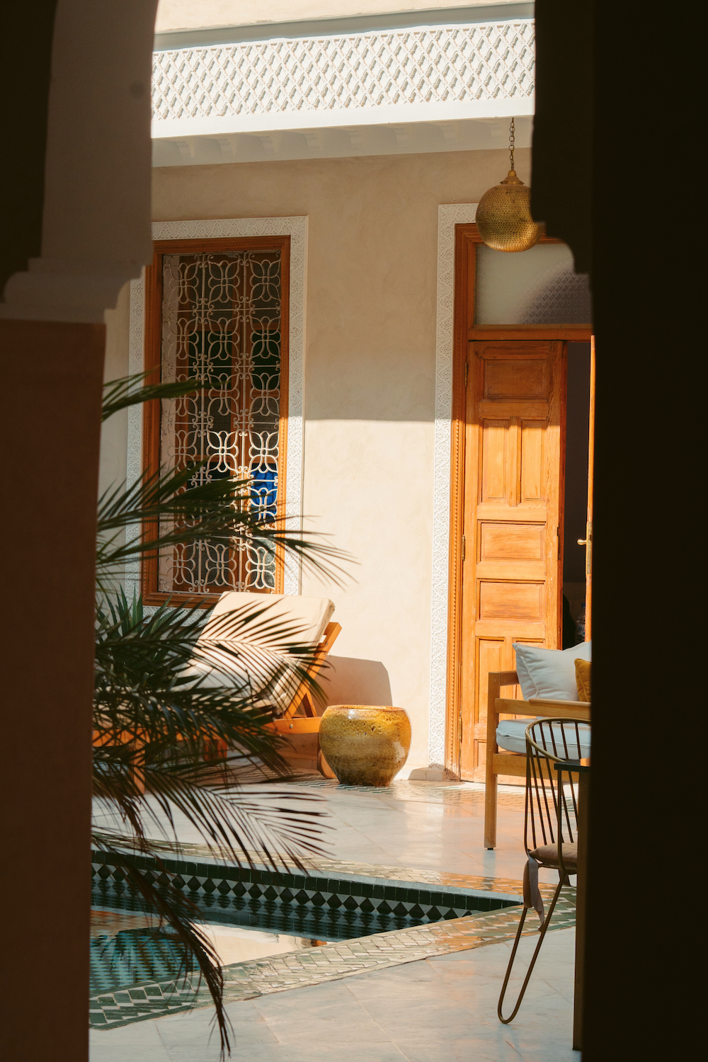 Interior view of a sunny courtyard with wooden door, lounge chair with cushions, potted plant, and pool, seen through a gap in the wall.