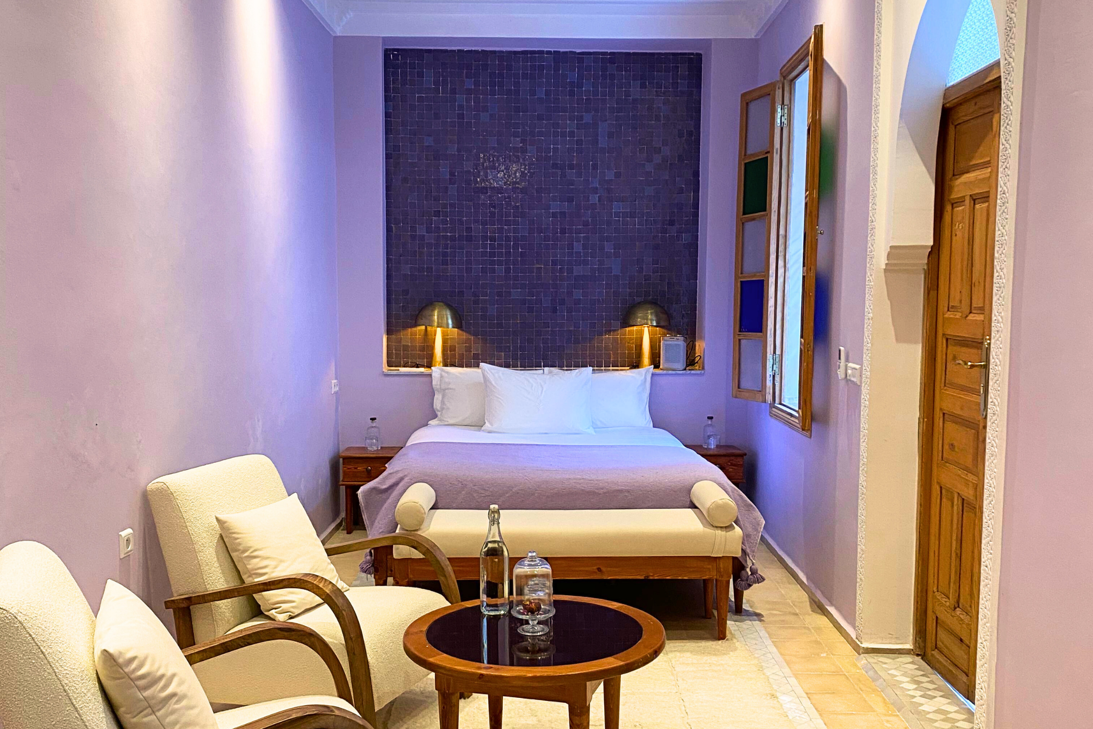 A hotel room with a bed, purple walls, and a dark tiled headboard. There are two wall-mounted lamps on either side of the bed, wooden nightstands, and open wooden shutters on the window. A cream-colored armchair and matching sofa with wooden armrests are positioned near a round wooden coffee table with a glass top, holding a water bottle and two glasses. The room has a cream rug on the tile floor and a decorative ceiling light fixture.