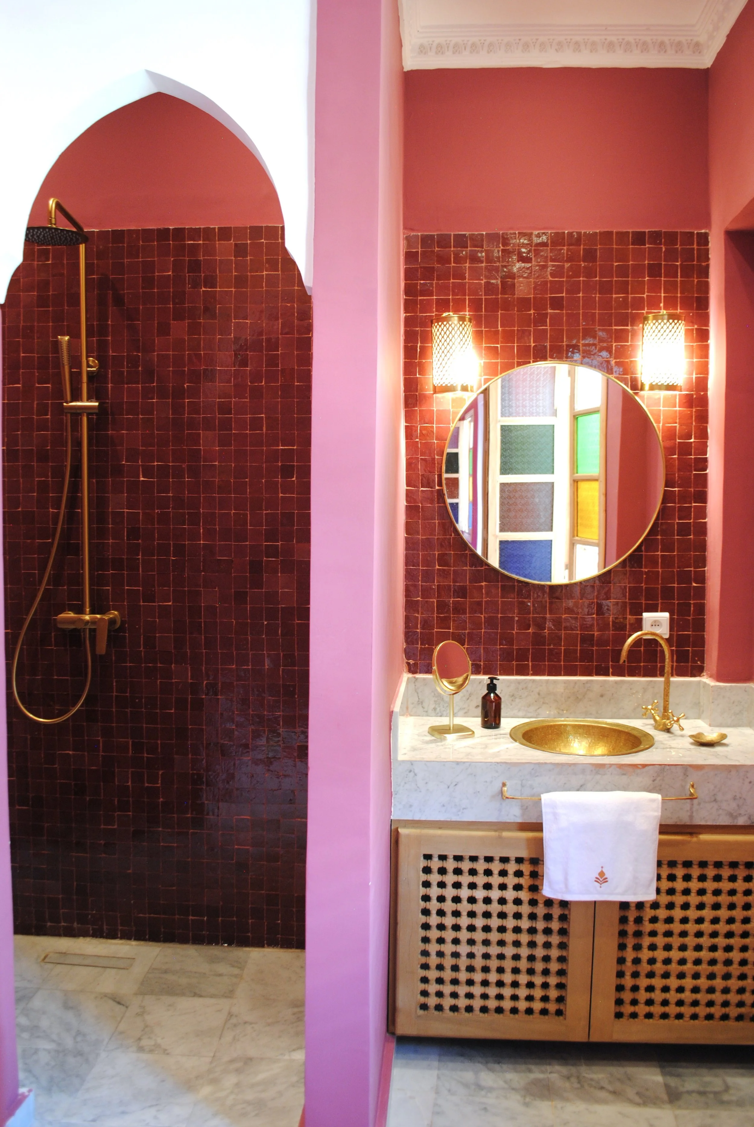 Bathroom with pink walls, a marble countertop with a gold sink, gold faucet, and mirror. There is a shower area with dark red tiles and a gold showerhead.
