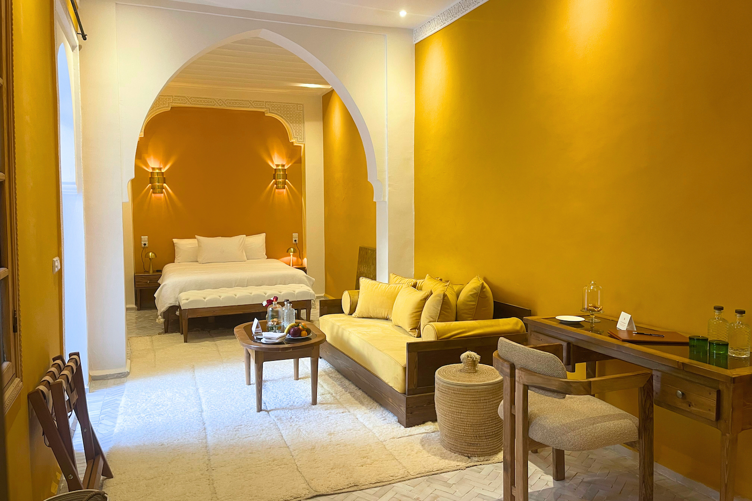 A cozy hotel room with a bed and a sitting area. The bed is in the background with white bedding and two pillows, illuminated by wall-mounted lamps. The sitting area in the foreground has a yellow sofa with matching cushions, a small wooden coffee table with bottles, a bowl of fruit, and a glass jar, and a wooden desk with a chair, bottles, and glasses. The room has warm yellow walls, arched architectural details, and soft lighting.