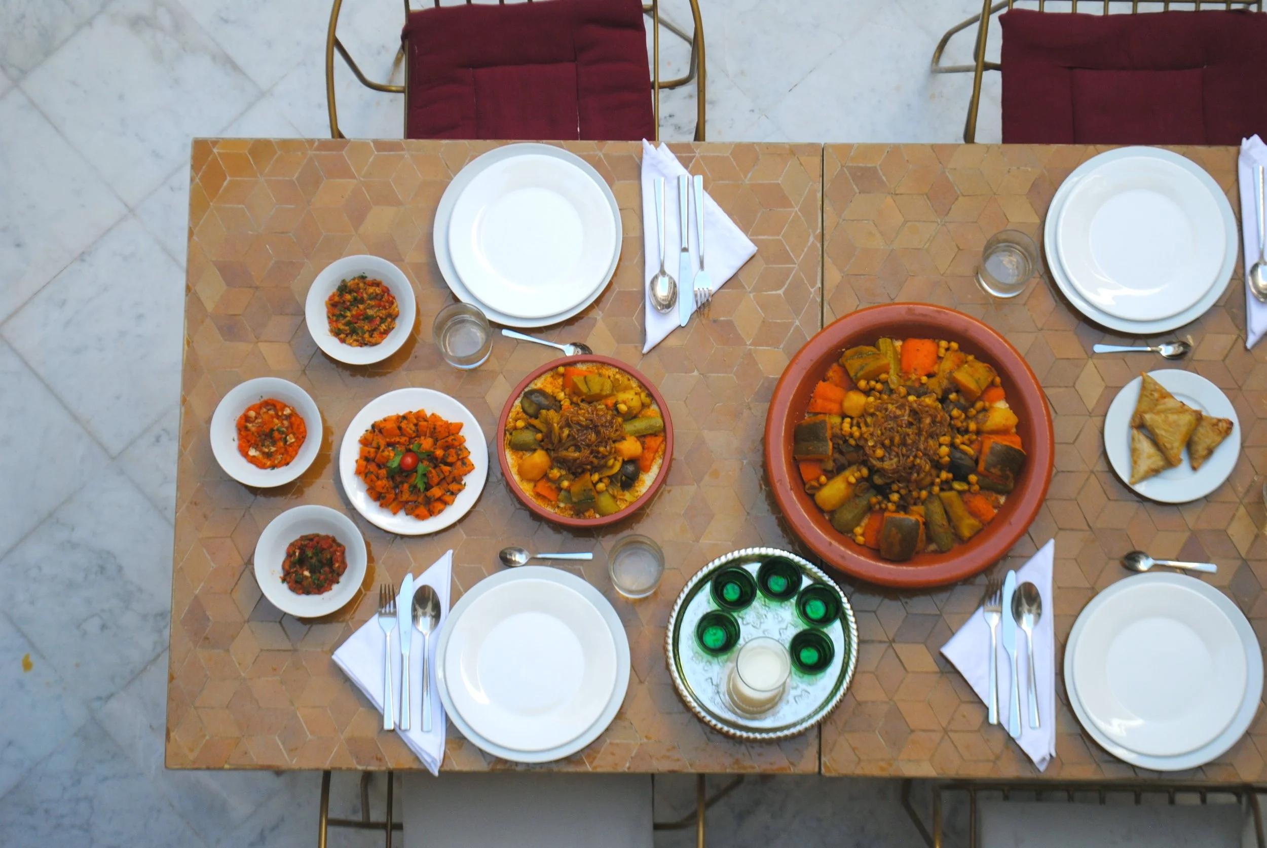 Table set with plates, silverware, glasses, and traditional Middle Eastern or North African dishes including a large tagine with vegetables and spices, smaller bowls of various cooked vegetables and dips, a tray with small glasses of tea, and a plate of pastries. The table has a geometric patterned surface and is surrounded by red-cushioned chairs.