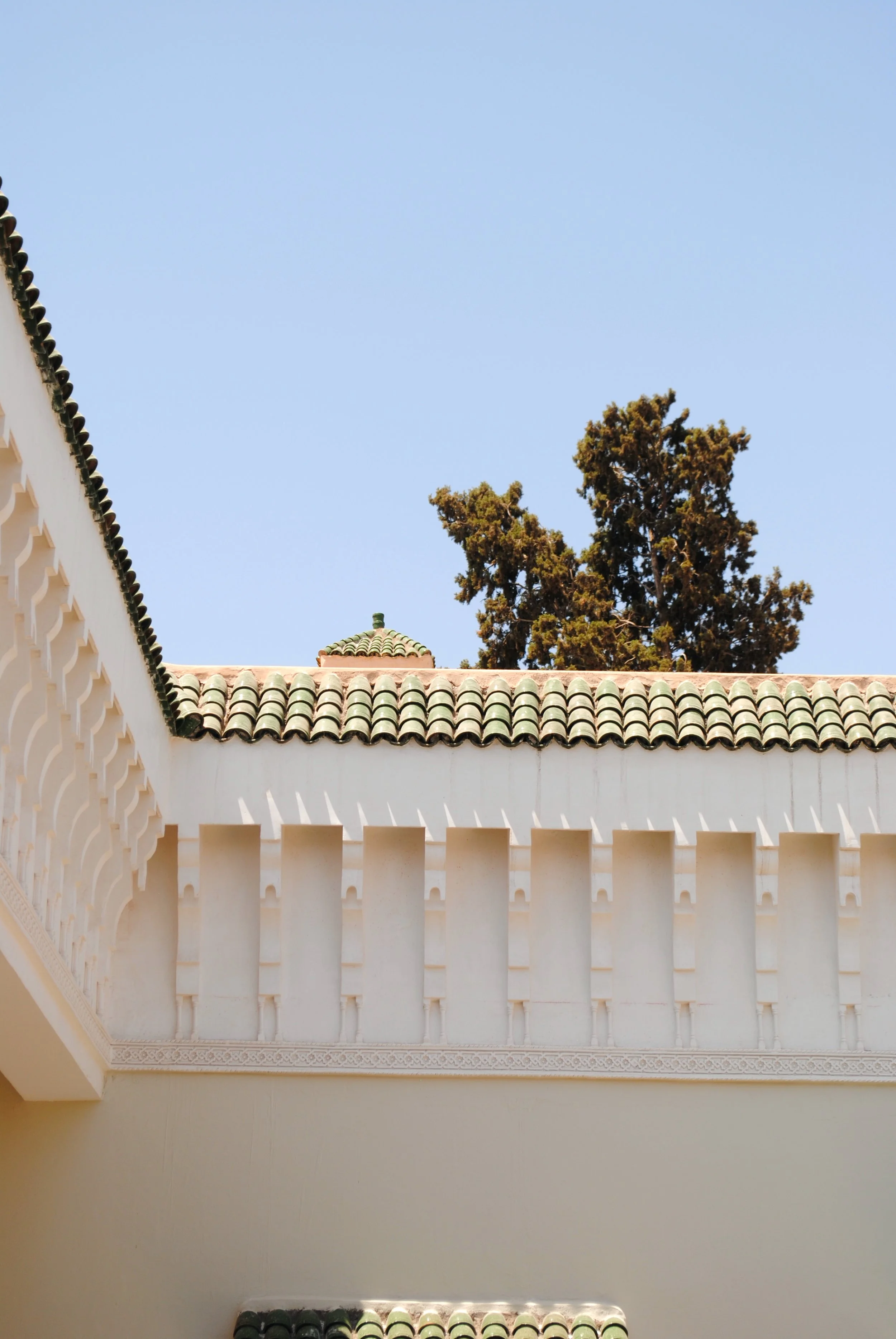 A view of a building with white walls, tiled roof, and decorative architectural elements, with a tall tree in the background under a clear blue sky.