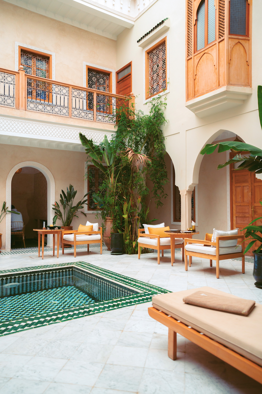 Interior courtyard with a small pool, sitting area with wooden chairs and cushions, potted plants, and ornate windows in a light-colored building with arched doorways.