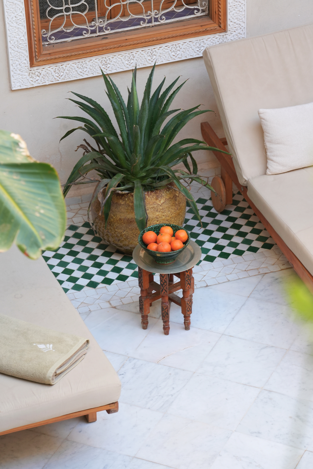 A large potted pineapple plant sits behind a small wooden table holding a bowl of oranges, with a beige cushioned sofa with a white pillow nearby. The floor is tiled with white and green patterned tiles, and the wall features a decorative white carved frame around a window with white iron bars.