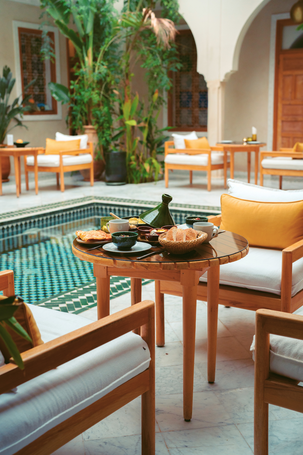 Indoor courtyard with a small pool surrounded by wooden furniture and lush greenery, set for breakfast with cups, plates, bread, and a green teapot on the table.