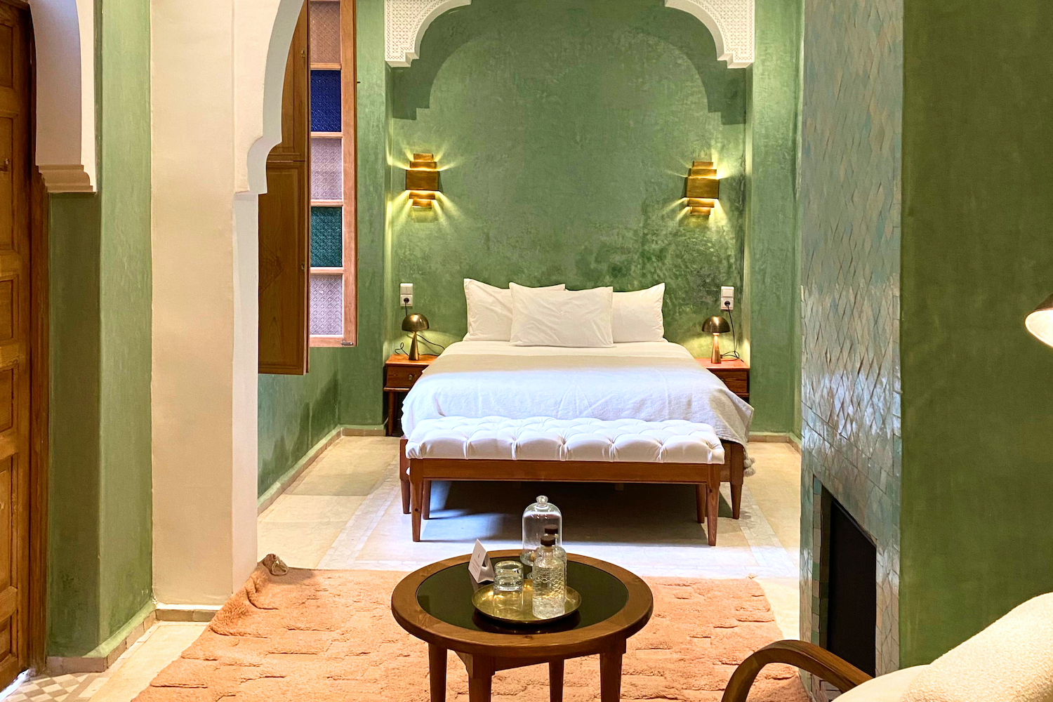 A cozy hotel room with green walls, a large bed with white linens, two bedside lamps, a white armchair, a small wooden table with a brass tray, and a pink rug.