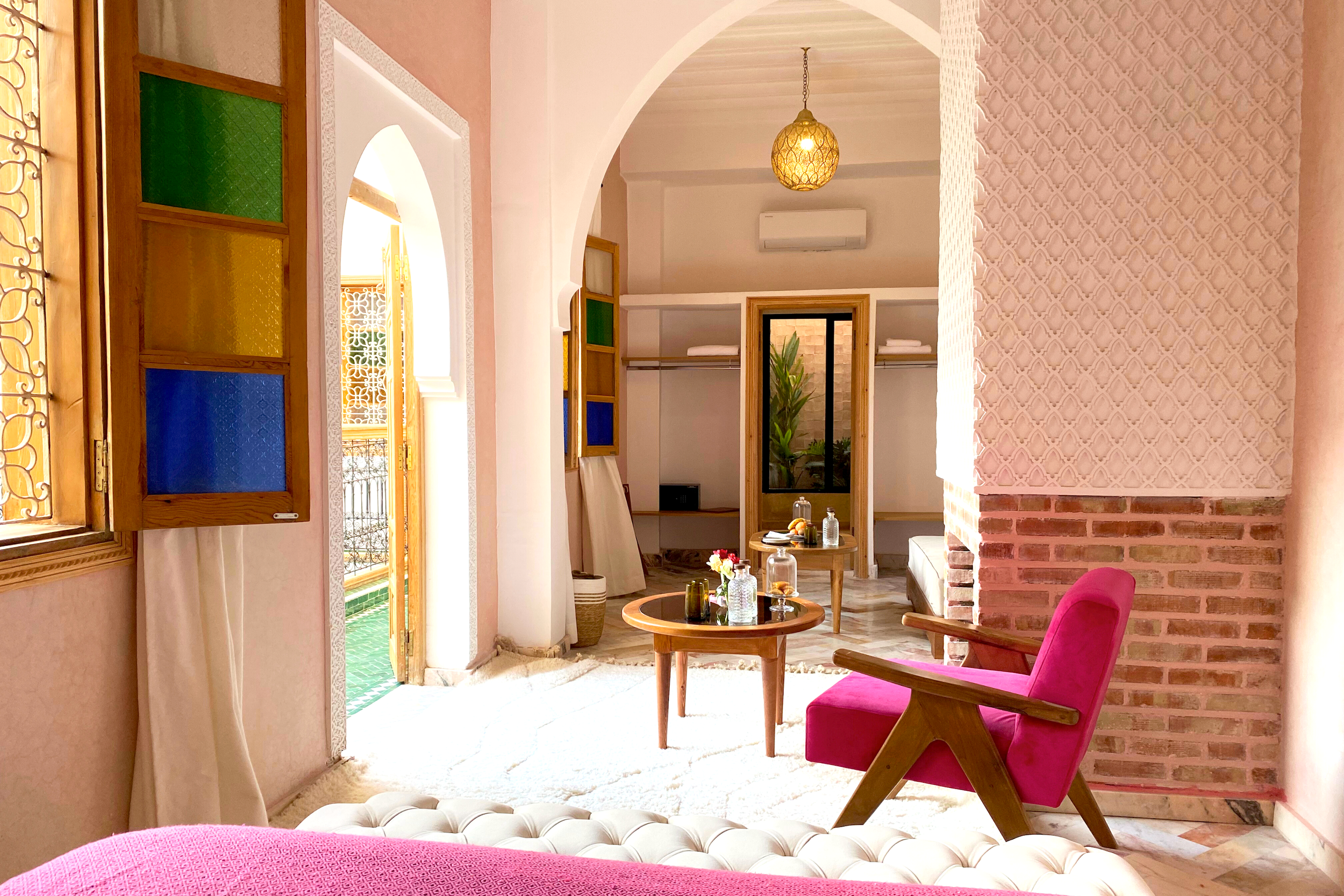 A cozy hotel room with green walls, a large bed with white linens, two bedside lamps, a white armchair, a small wooden table with a brass tray, and a pink rug.