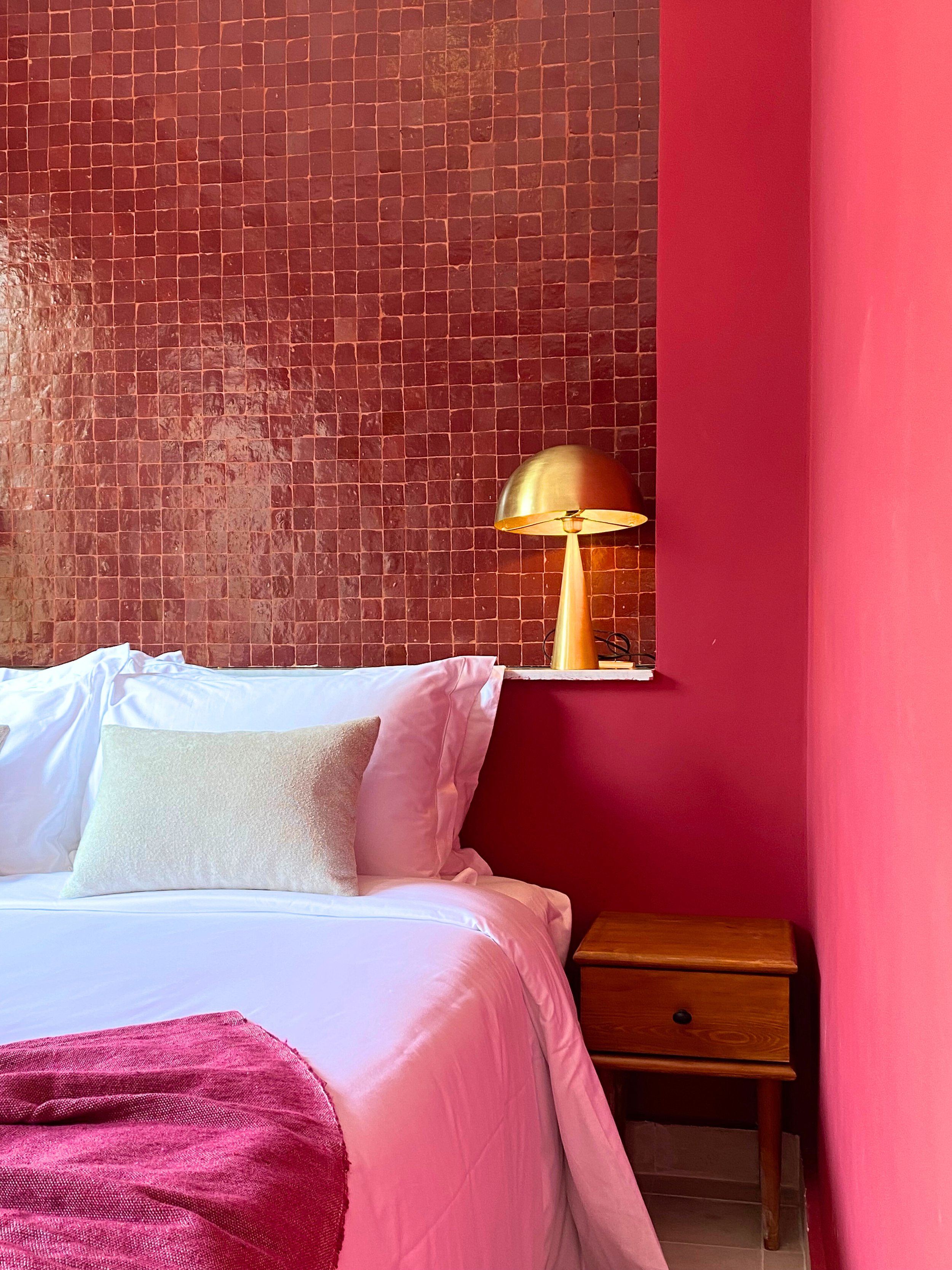 A corner of a bedroom with pink walls, a white bed with pillows, a wooden nightstand, and a gold bedside lamp against a pink tiled accent wall.