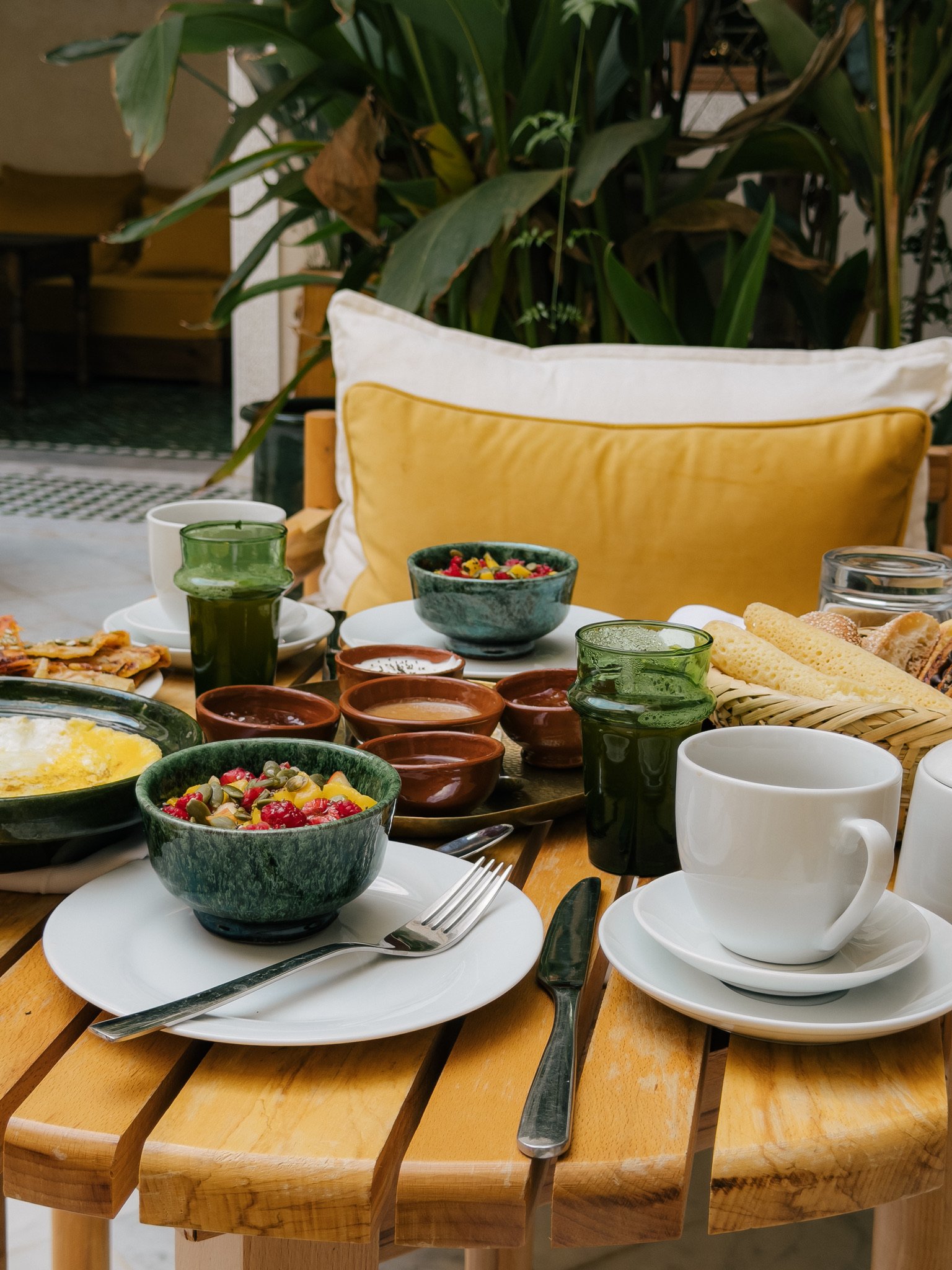 Breakfast table with bowls of fruit, mugs, glasses of green juice, bread, and small dishes on a wooden table outside.