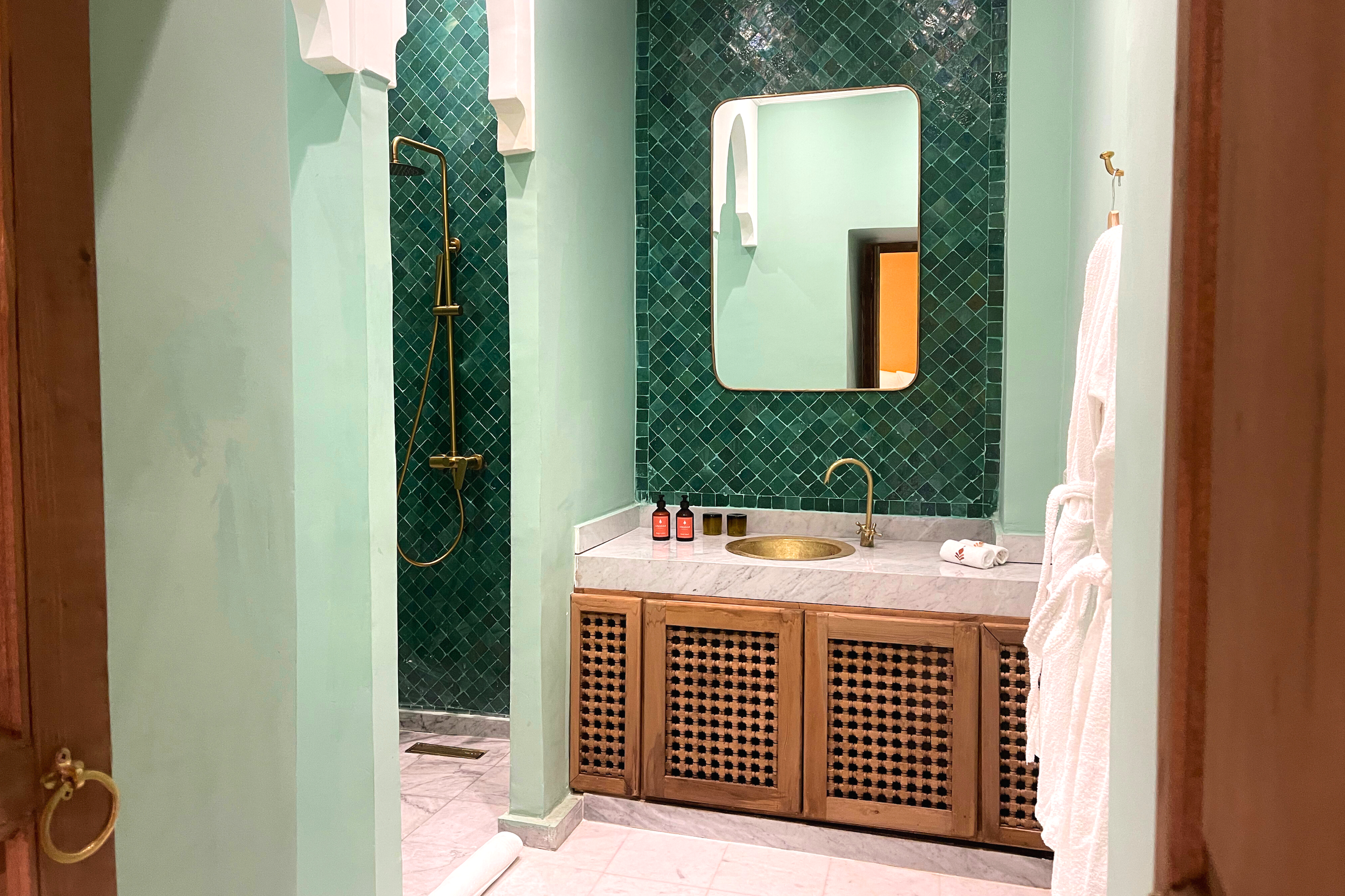 Bathroom with pink walls, a marble countertop with a gold sink, gold faucet, and mirror. There is a shower area with dark red tiles and a gold showerhead.