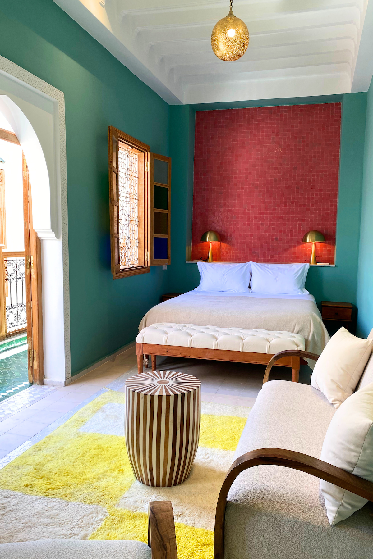 A bedroom with teal walls, a red textured accent wall behind the bed, a gold spherical ceiling light, two gold bedside lamps, a wooden window with stained glass, a white upholstered bench at the foot of the bed, a beige sofa with white pillows, a striped round ottoman, and a yellow and cream rug.