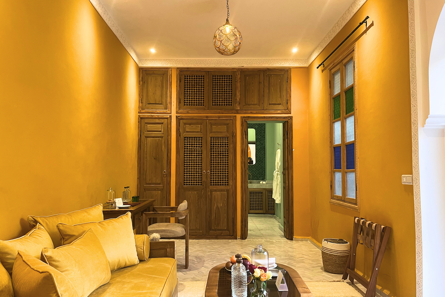 Living room with yellow walls, a yellow sofa with pillows, a coffee table with flowers and bottles, a wooden chair, and a window with colored panes, leading to a bathroom with green tiles.