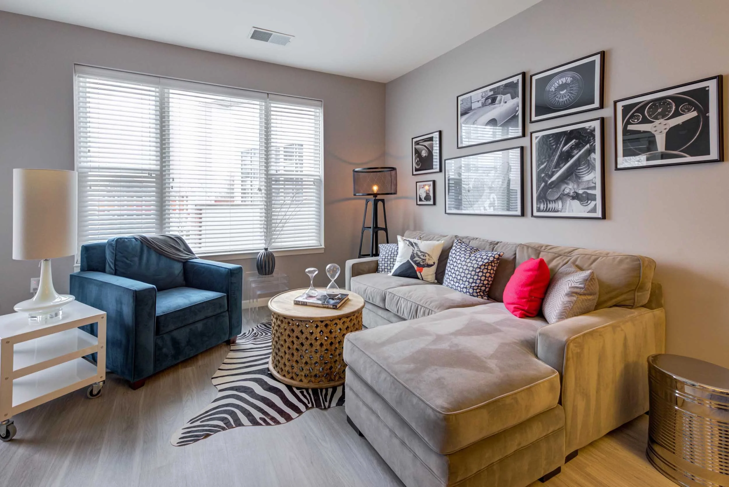 A living room with large window blinds, a beige sofa with cushions, a blue armchair, a zebra print rug, a round wicker coffee table, a white side table with a lamp, wall art featuring car parts, a floor lamp, and a decorative woven basket.