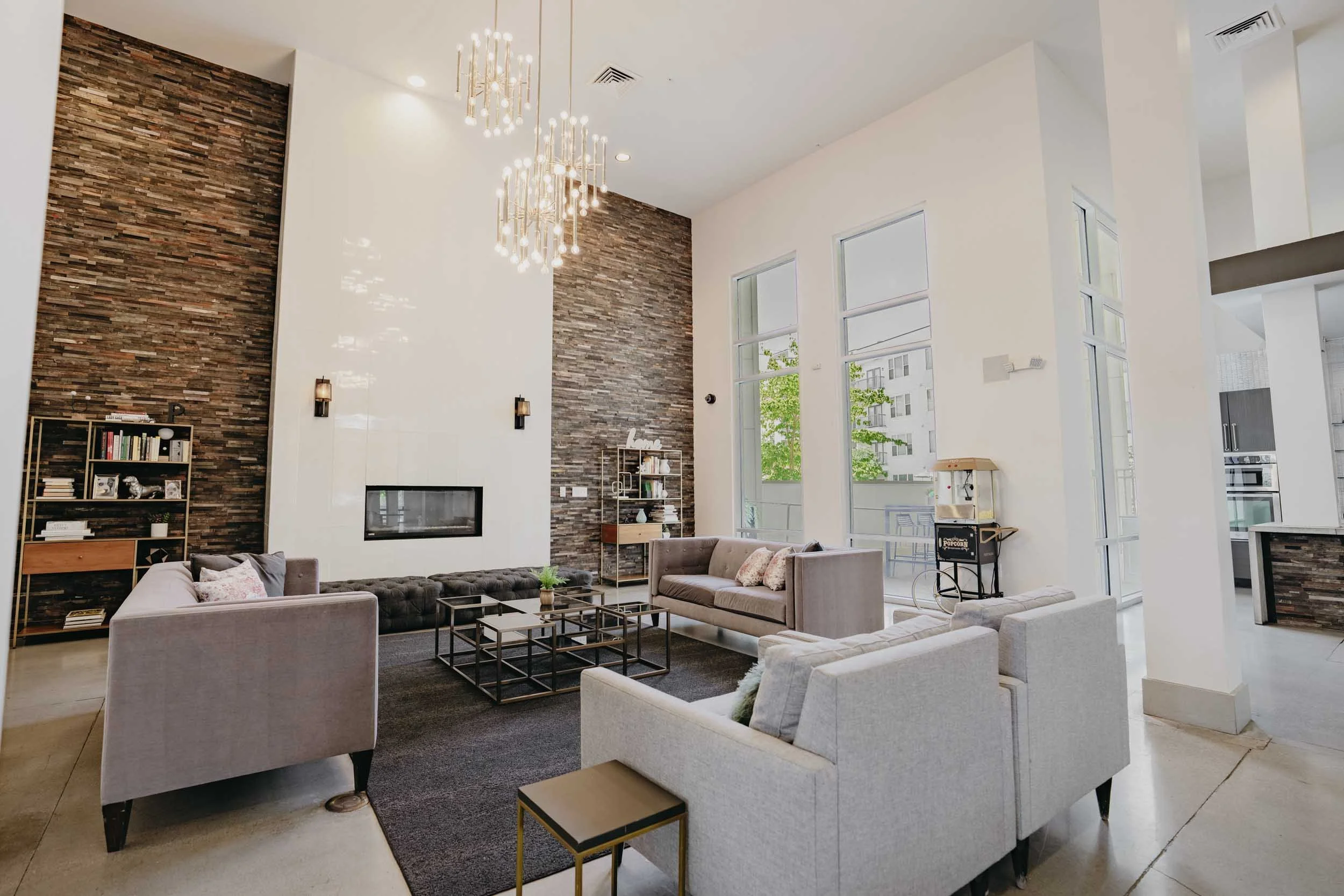Modern clubhouse room with high ceilings, large windows, and a mix of white and dark brown textured walls, furnished with multiple sofas, a black coffee table, and a bookshelf.