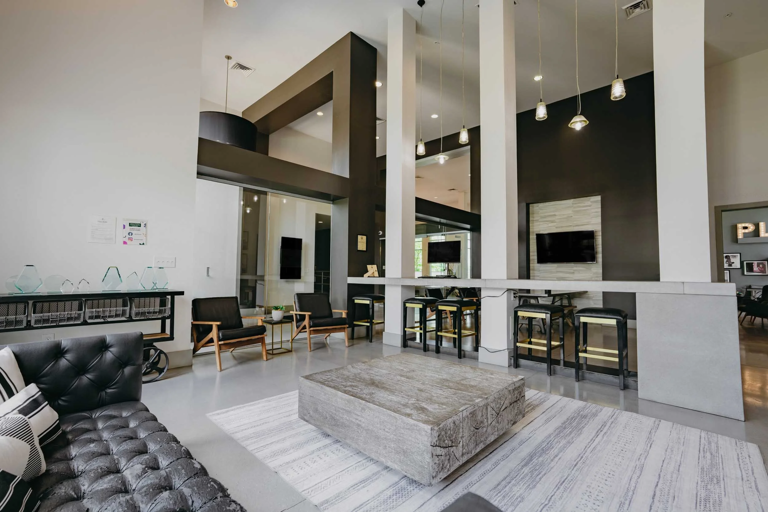 Modern lobby with black leather tufted sofa, wooden chairs, bar seating with black and yellow stools, pendant lights, and wall-mounted TVs.
