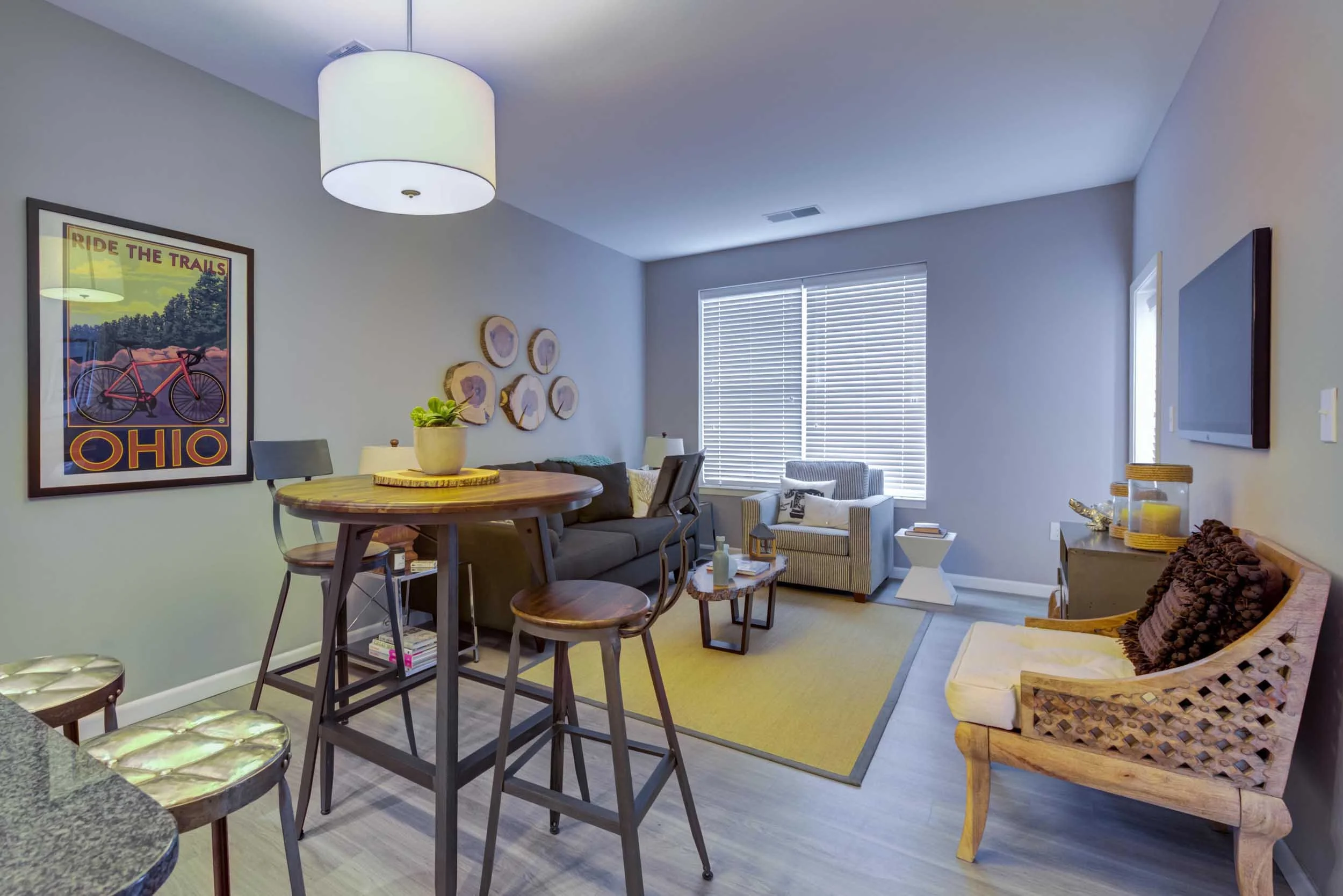 A cozy living room and dining area with modern and rustic decor, including a round wicker table, bar stools, armchairs, wall art, and a window with blinds.
