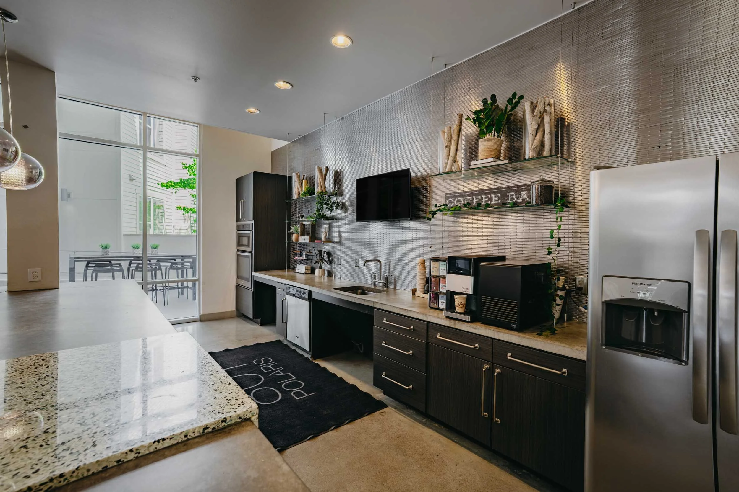 Modern kitchen with black cabinets, a light-colored speckled granite counter, and a backsplash with small metallic tiles. There are plants and decorative items on glass shelves, a small black TV, and a refrigerator. Natural light comes through large 
