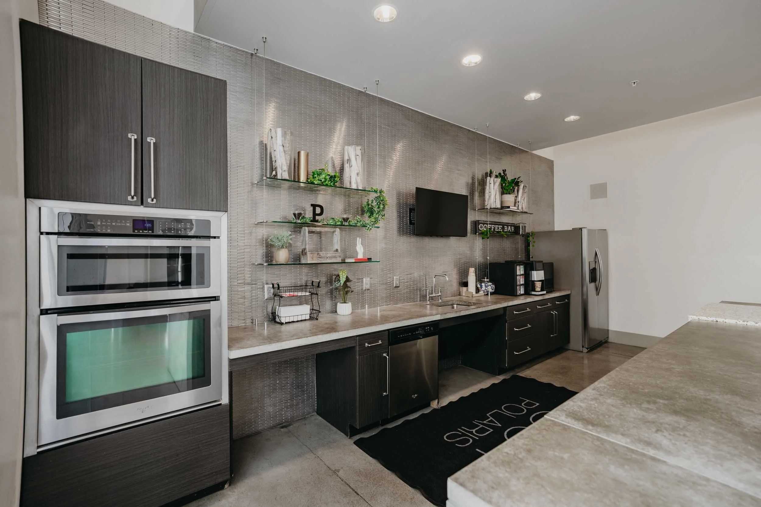 Modern kitchen with gray cabinets, stainless steel oven and refrigerator, black lower cabinets, beige countertops, open shelving with decorative items, a black rug with white text, and potted plants.