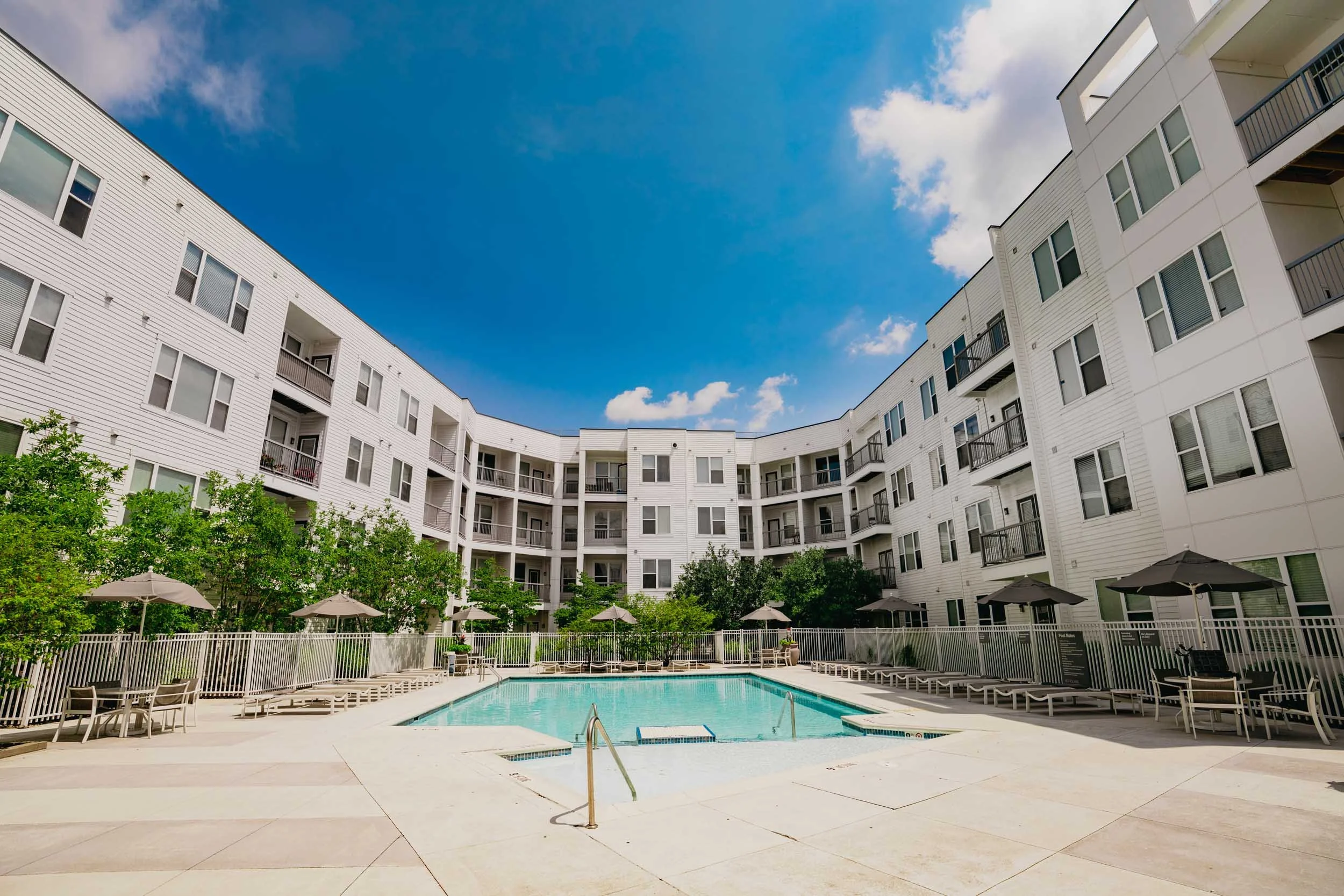 An outdoor swimming pool area within a residential complex with white apartment buildings surrounding it, green trees, and several patio umbrellas.