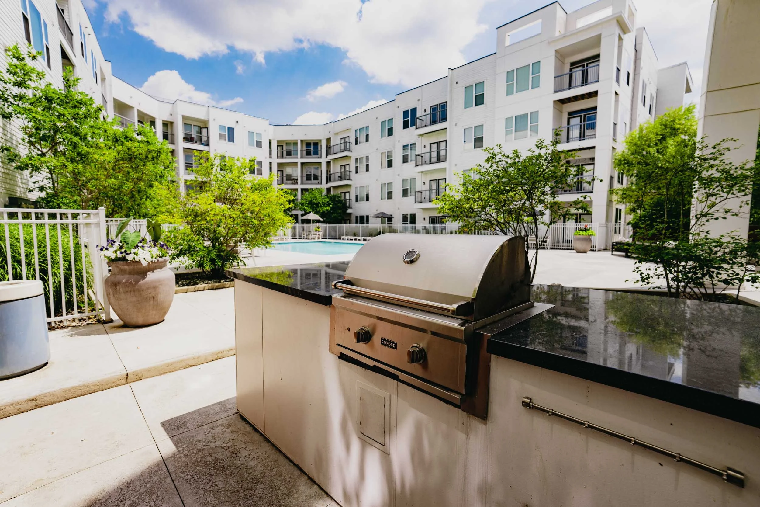Outdoor patio area of an apartment complex with a grill, trees, and a swimming pool surrounded by an apartment building with multiple balconies.