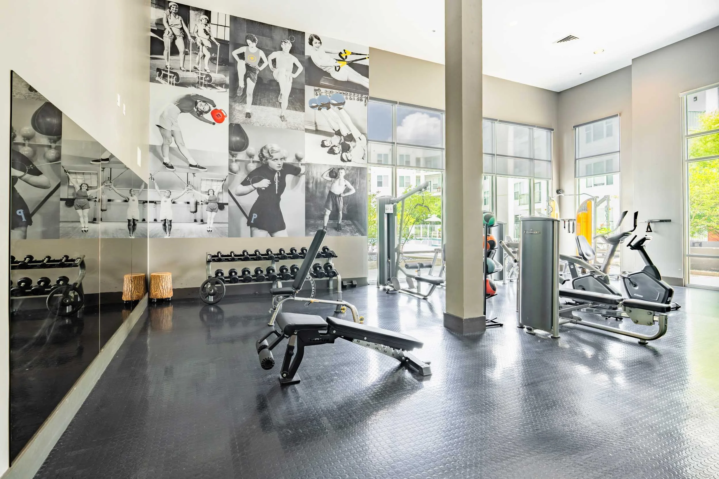 Modern gym interior with workout equipment, large windows, and a black-and-white vintage fitness mural on the wall.