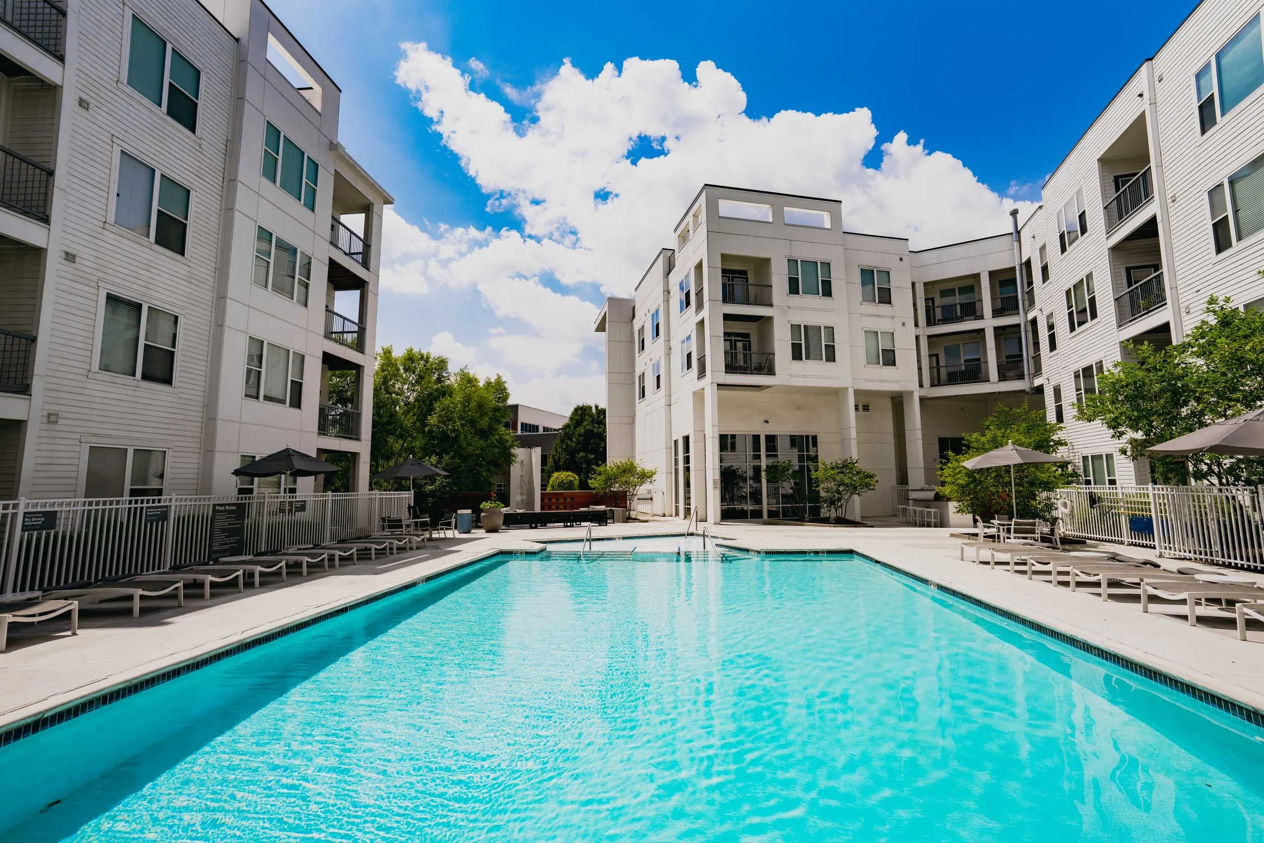 Apartment complex with a swimming pool in the courtyard, surrounded by white multi-story buildings under a blue sky with scattered clouds.