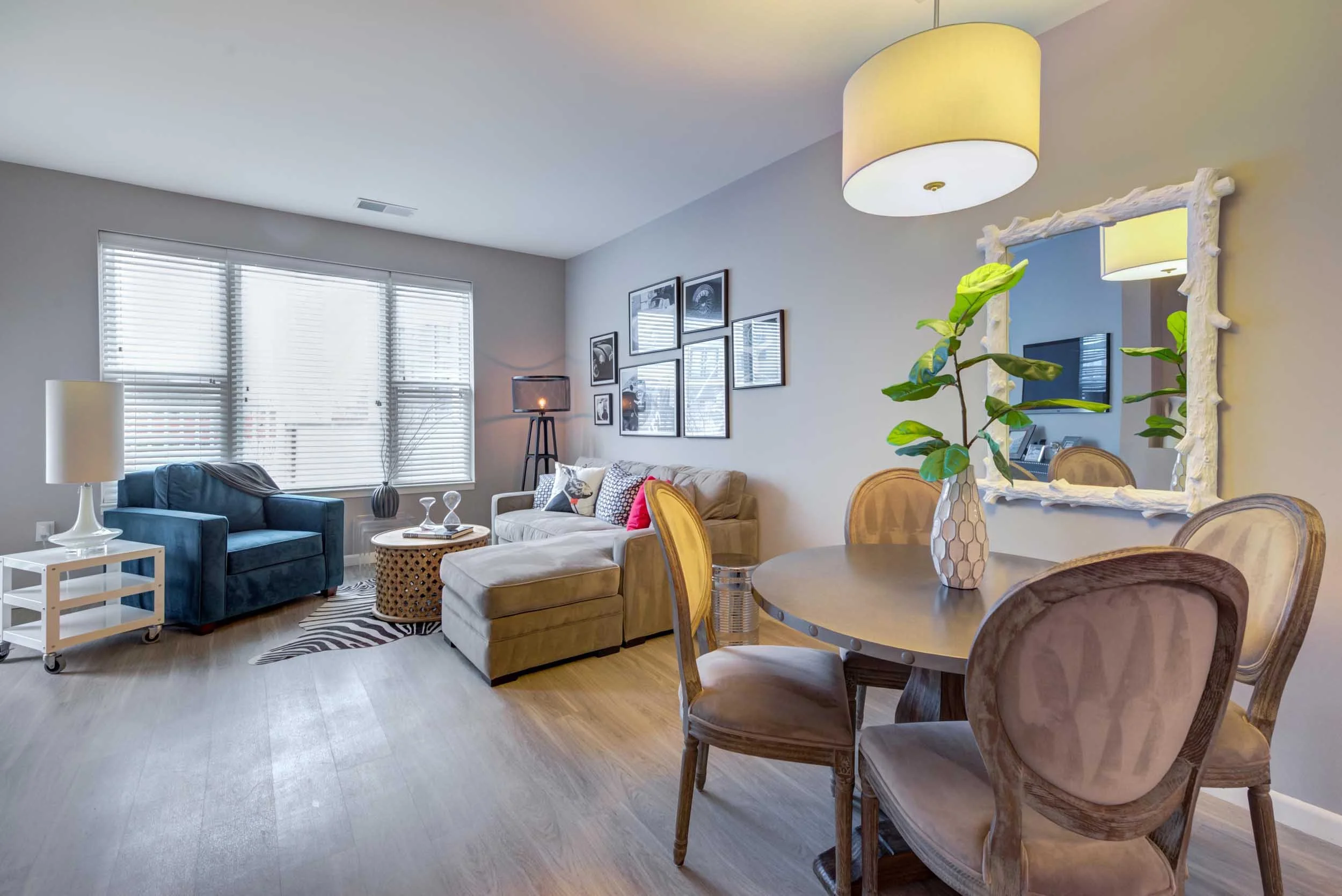 Living room with large window blinds, a blue armchair, a beige sofa, a round coffee table with decor, a zebra print rug, wall art, a black floor lamp, and a dining table with wooden chairs and a vase with greenery.