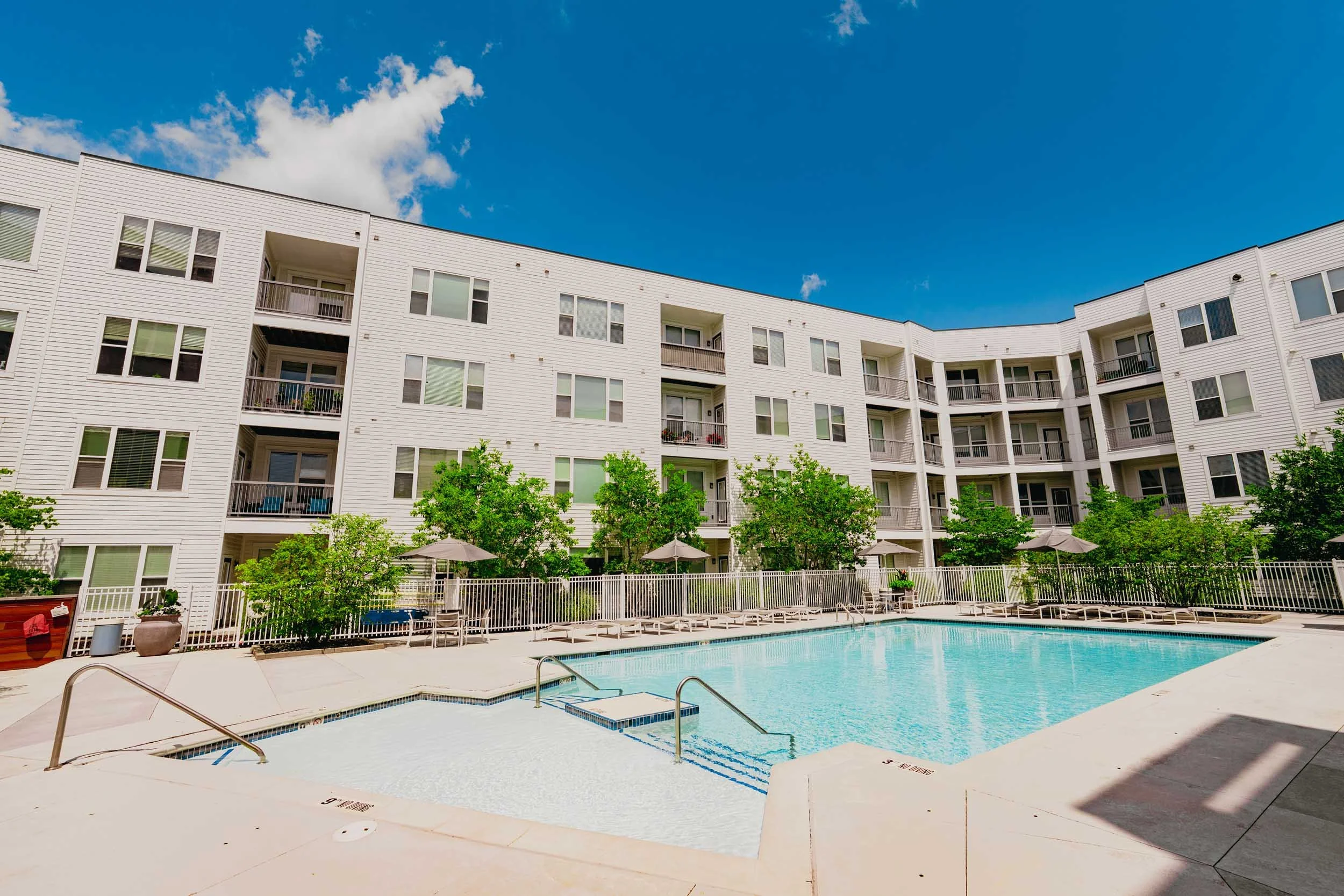 An outdoor swimming pool surrounded by a white apartment complex with multiple balconies under a bright blue sky with some clouds and green trees around the pool.