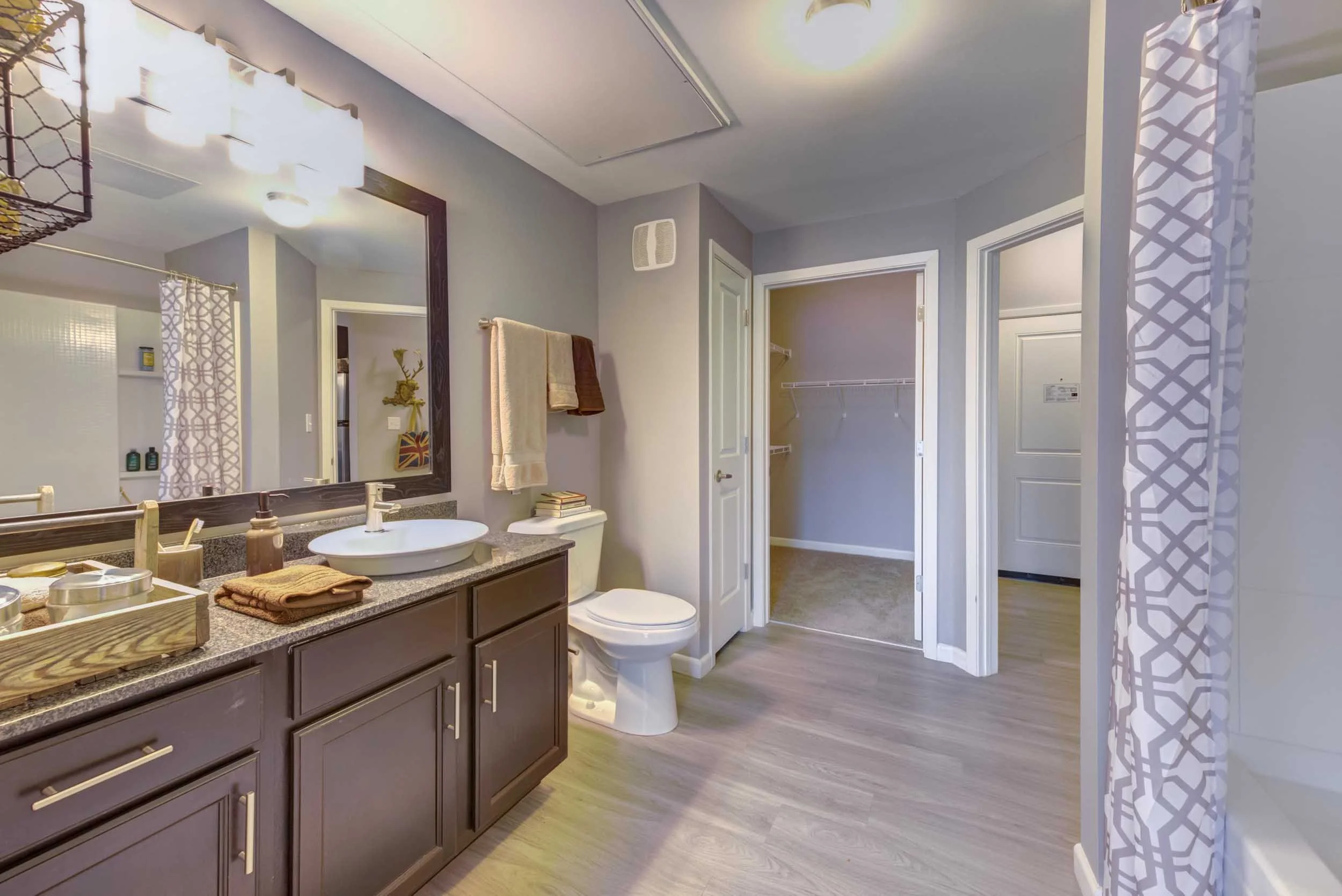A bathroom with a dark wooden vanity, granite countertop, white vessel sink, and various toiletries. A large mirror with a dark frame and wall-mounted towel rack with towels are above the vanity. To the right, there is an open doorway leading to a wa