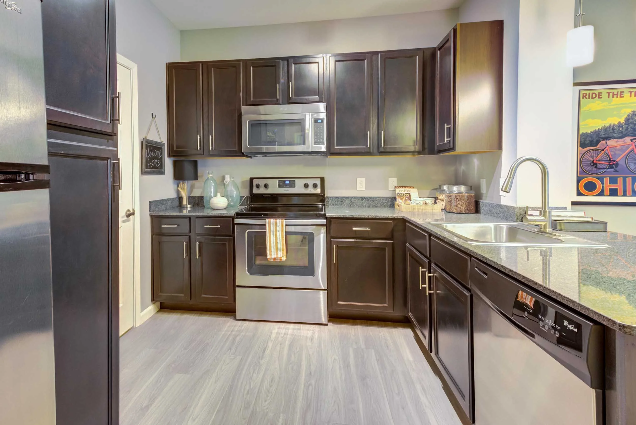 A modern kitchen with dark brown cabinets, stainless steel appliances including an oven, microwave, and a refrigerator. There are decorative vases and kitchen items on the countertops, and a colorful poster on the wall featuring a bicycle and the wor