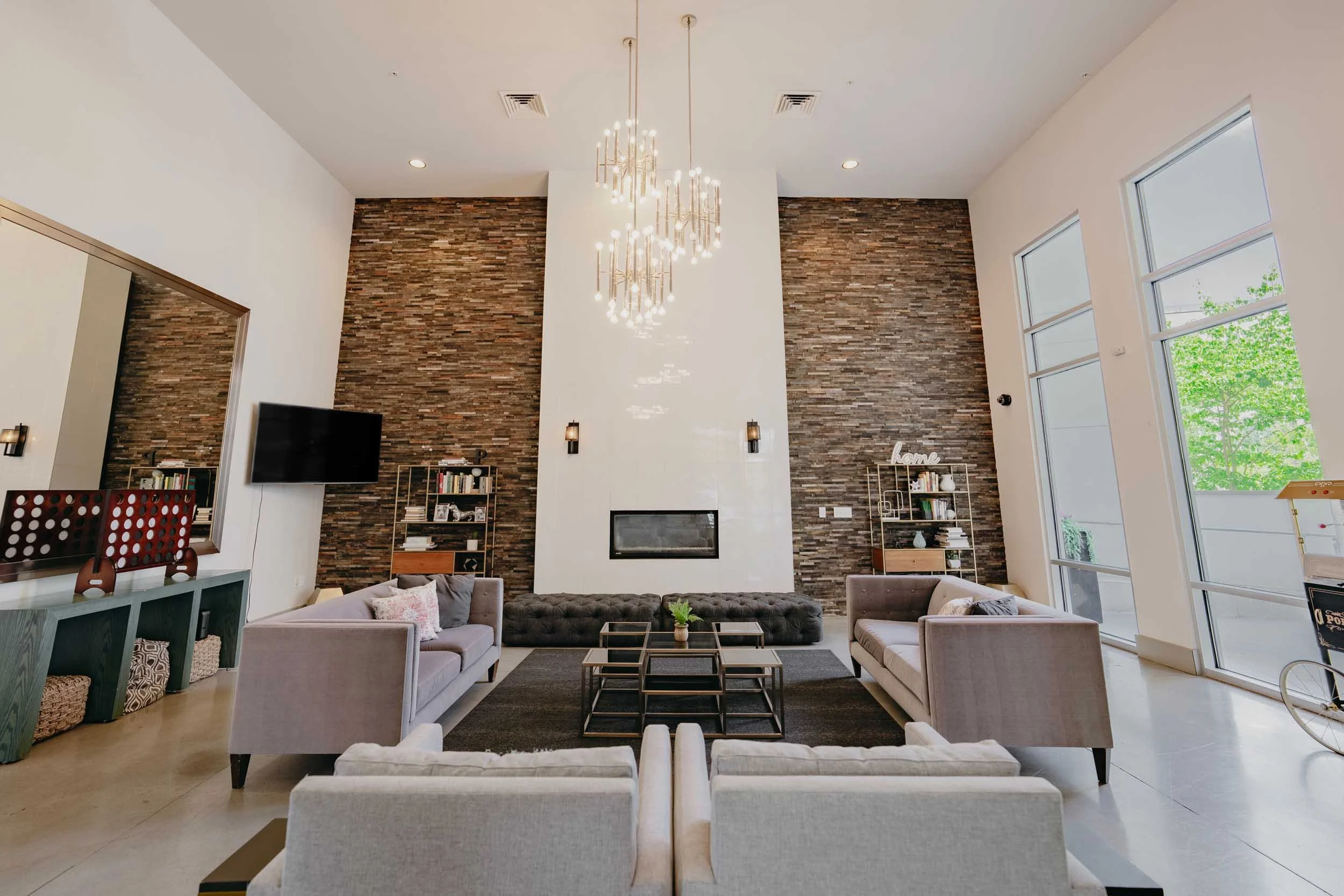 Modern club room with high ceiling, large windows, and a tall brick accent wall. Features include two light-colored sofas, a dark tufted ottoman, a coffee table, and built-in bookshelves.