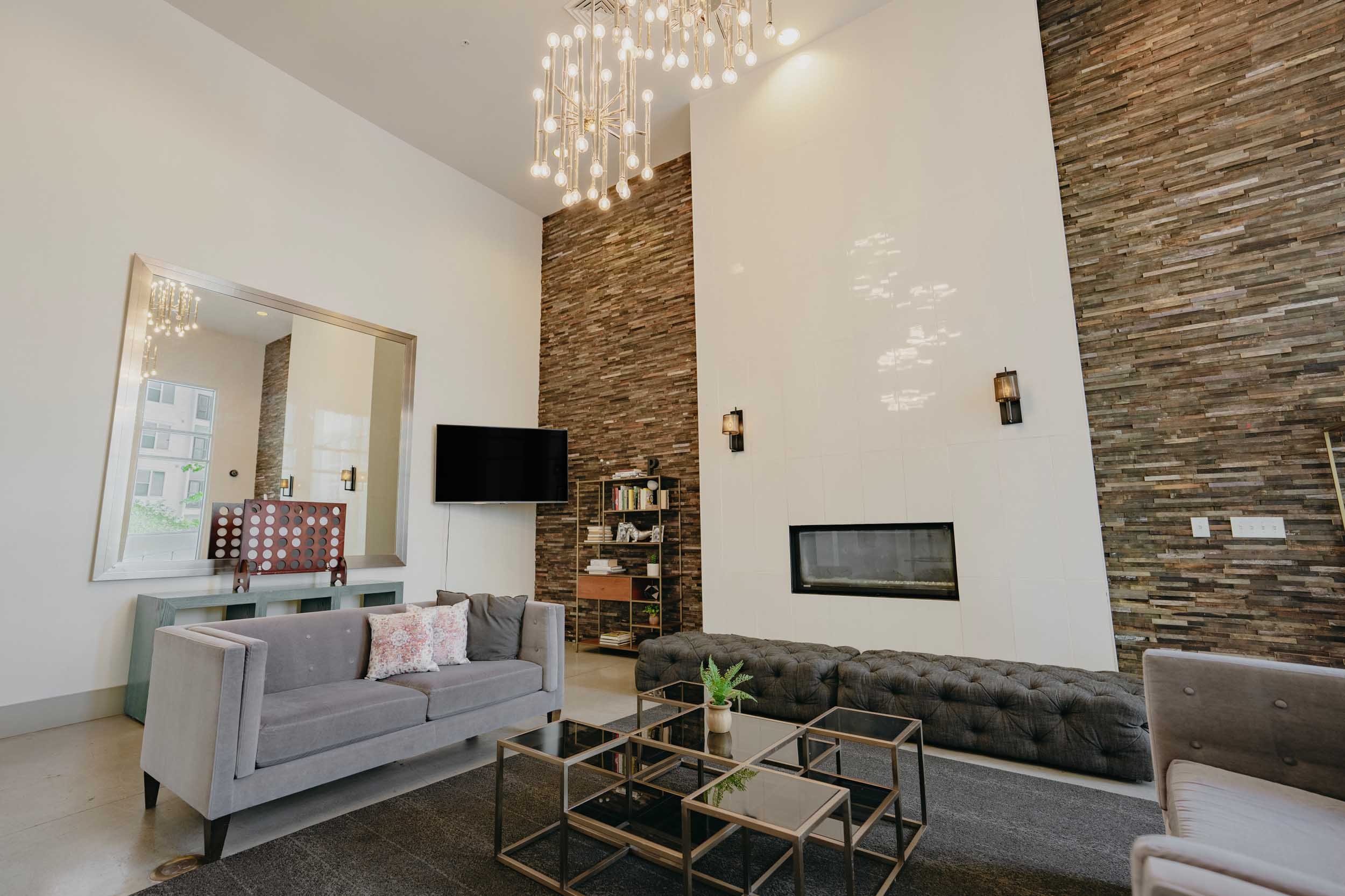 Modern club room with a gray sofa, glass tables, a textured stone accent wall, wall-mounted lights, a large mirror, a flat-screen TV, and a chandelier with multiple bulbs.