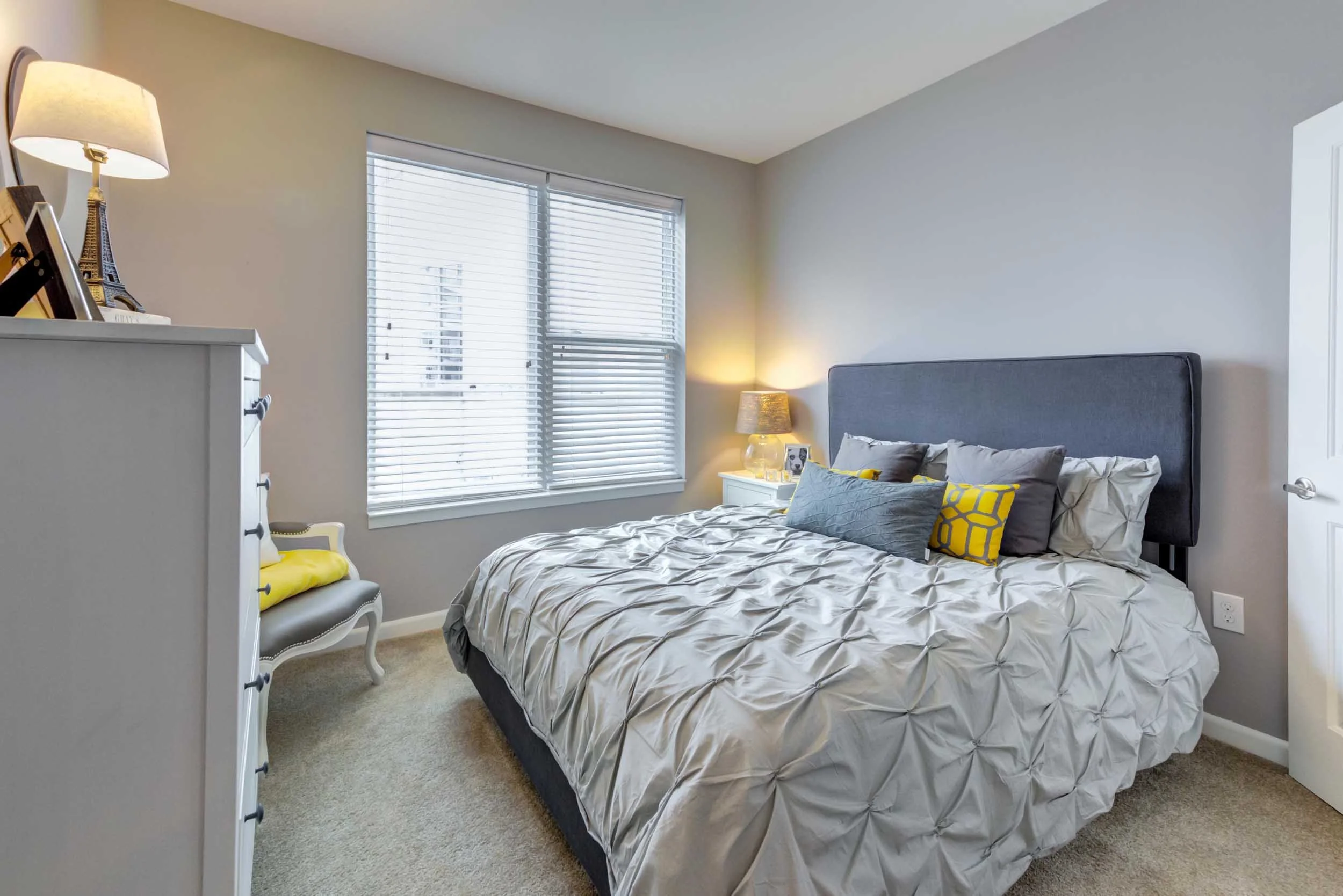 A bedroom with a large bed, a gray upholstered headboard, and multiple pillows. There are two bedside tables with lamps and a pair of pictures on the bedside table. A dresser with decorative items and a lamp is nearby. The room has light-colored wall