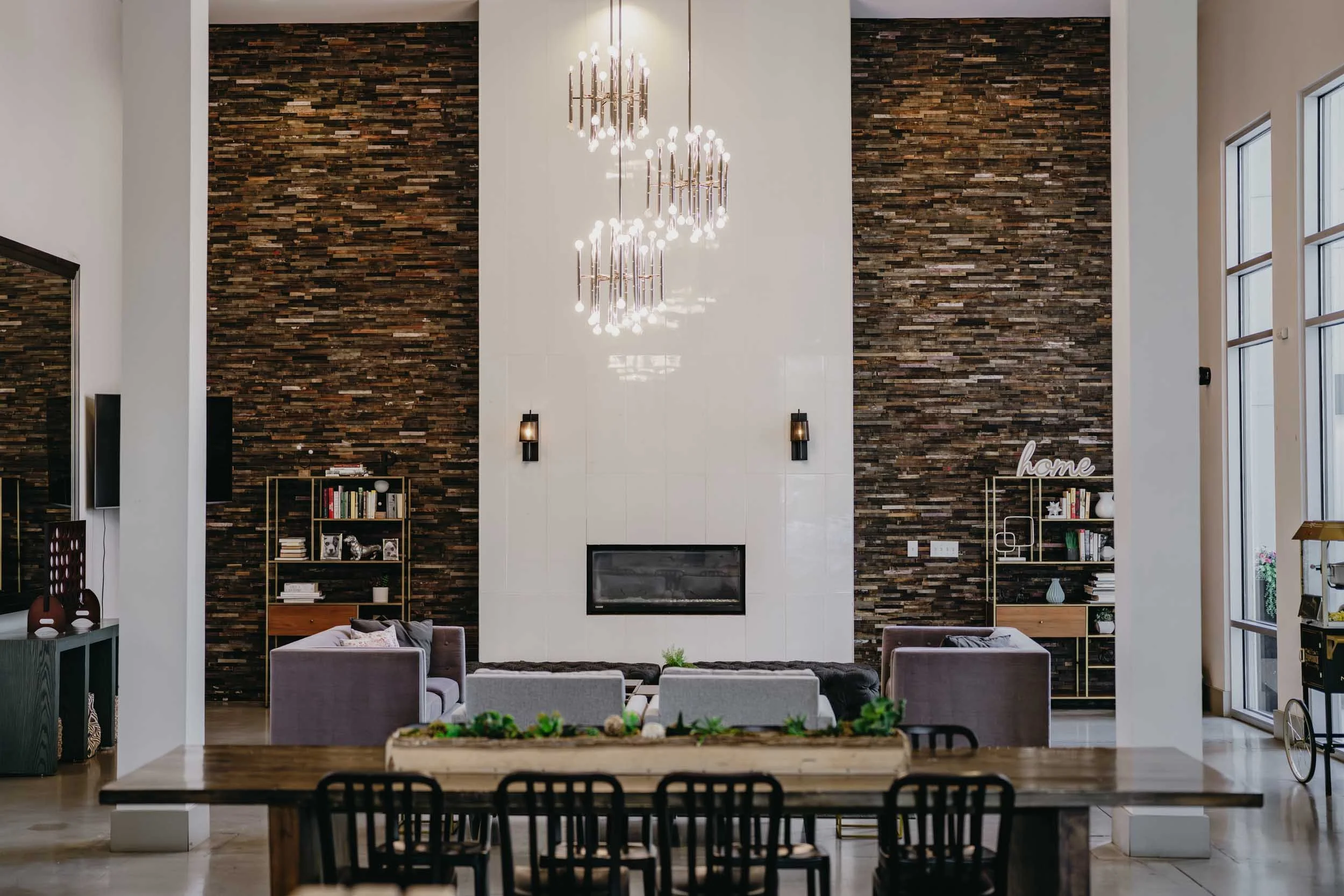 Bright living room with modern decor, featuring a large white fireplace, dark wooden accent walls, high windows, and a chandelier hanging from the ceiling.