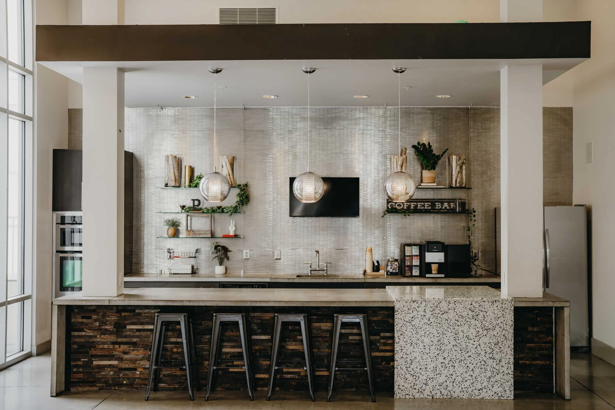 Modern kitchen with a granite countertop island, black bar chairs, pendant lights, a textured backsplash, plants, books, and coffee-related decor.