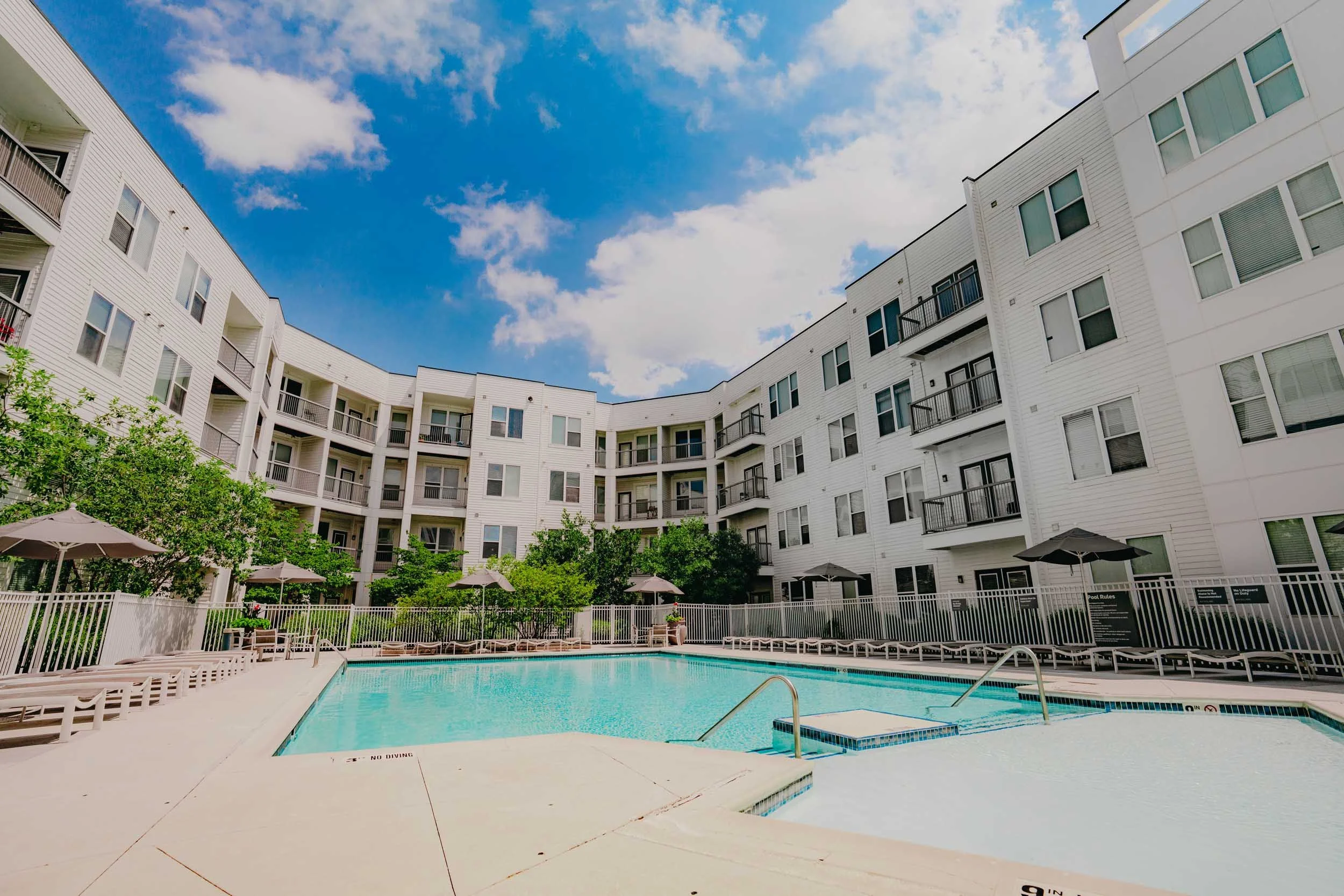 An outdoor swimming pool area surrounded by white apartment building with balconies, green trees, and blue sky with scattered clouds.