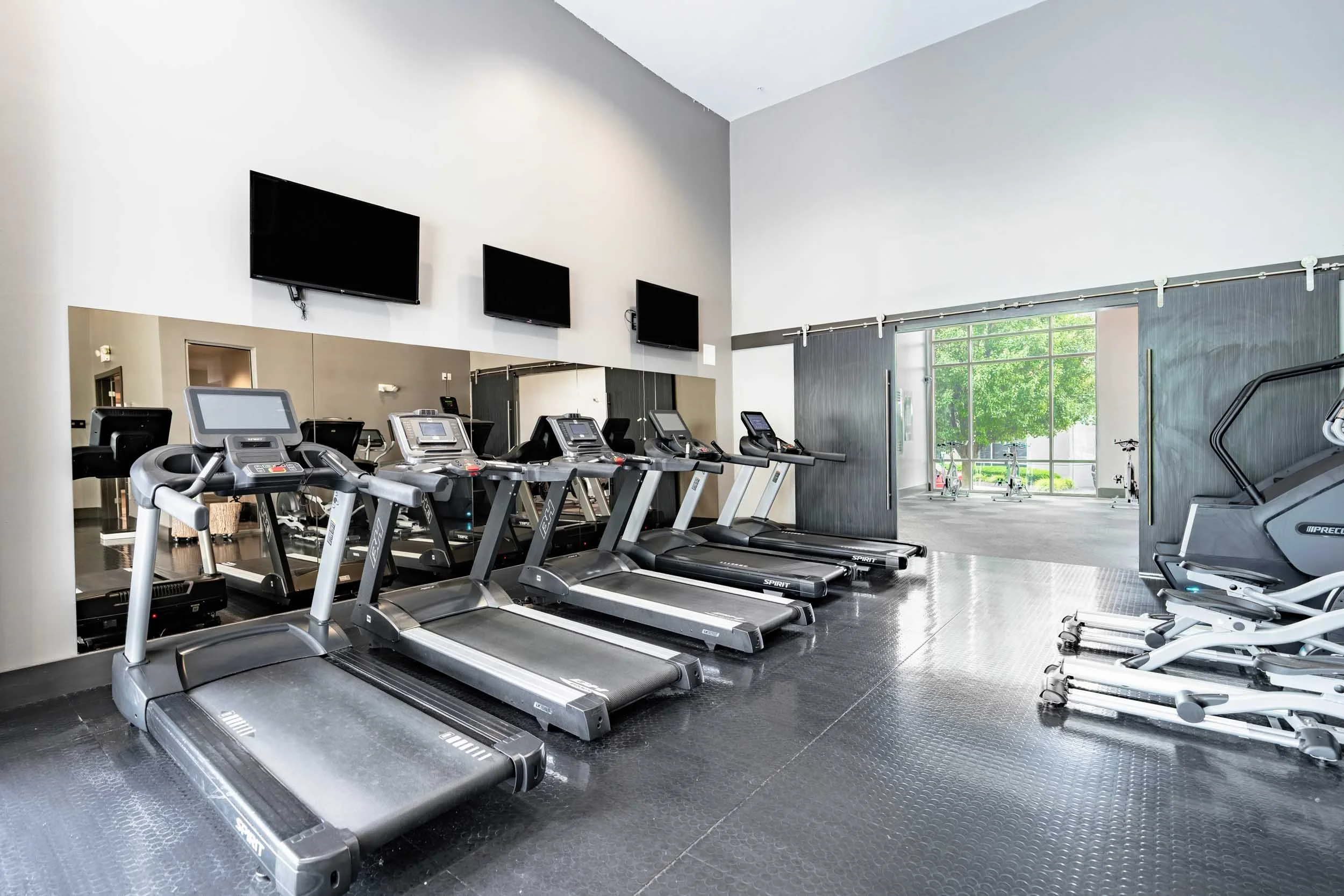 Empty gym with multiple treadmills and other exercise equipment near a large window showing greenery outside.
