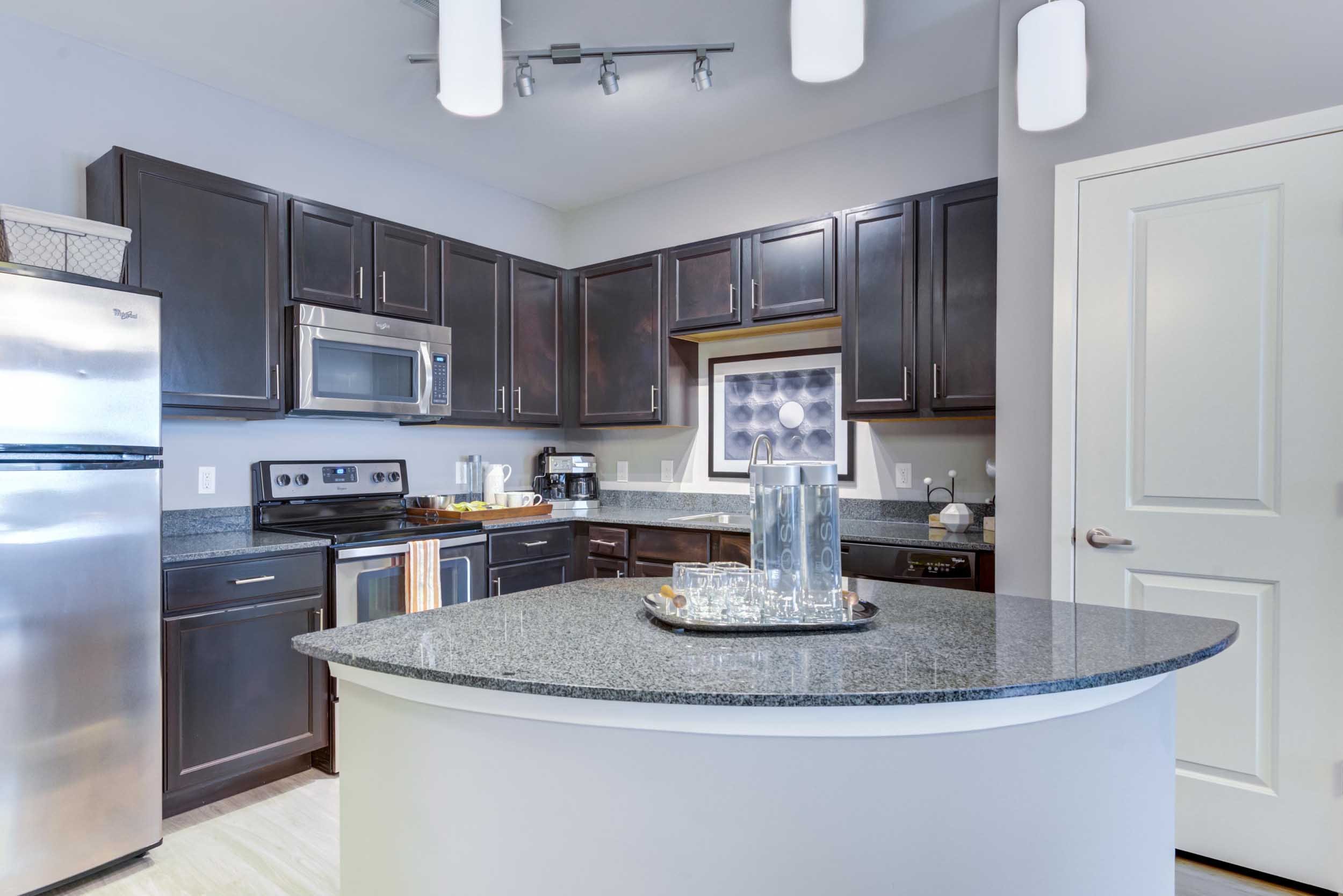 Modern kitchen with dark wood cabinets, stainless steel appliances including a microwave and refrigerator, a granite countertop, a small window, and track lighting on the ceiling. A tray with glasses and water bottles is on the island, and a white door is visible on the right.