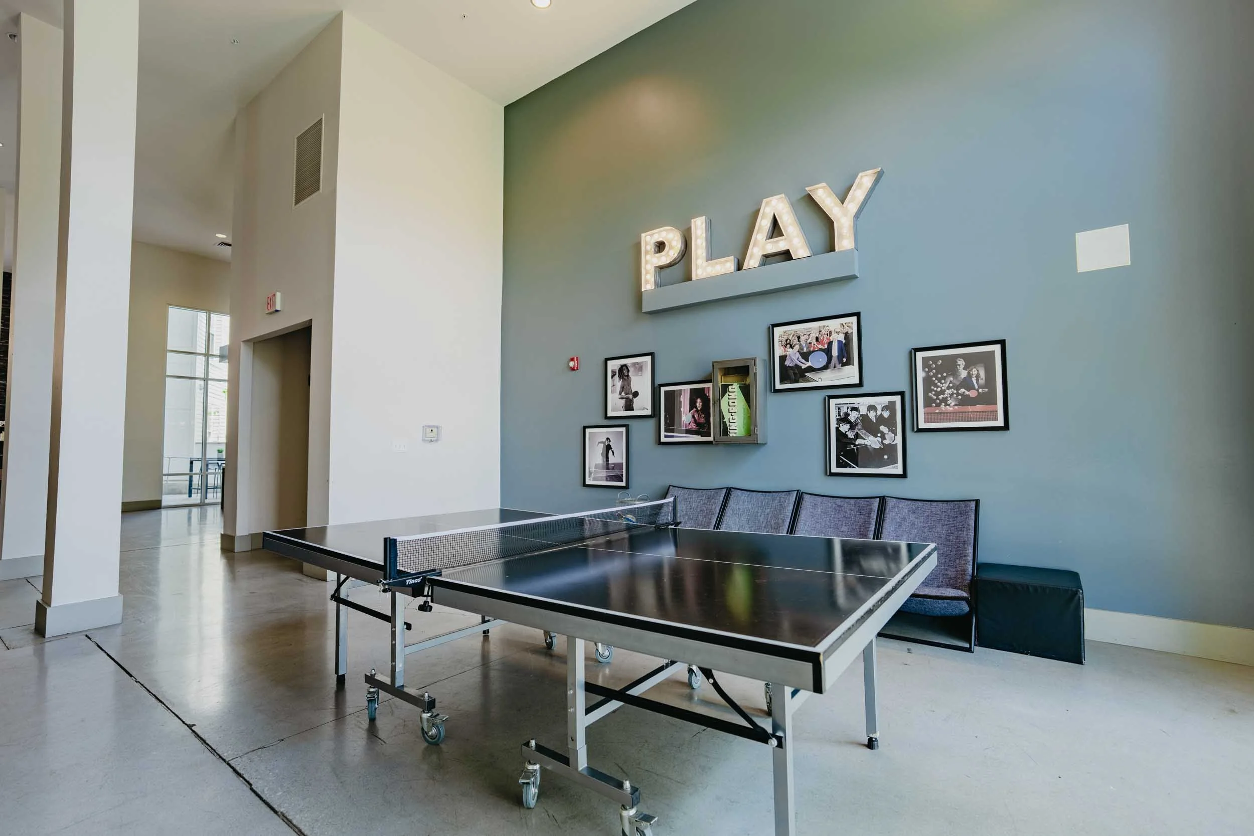 An indoor recreational area with a ping pong table and a seating area against a light blue wall. The wall features framed photos and a large illuminated sign spelling 'PLAY'.