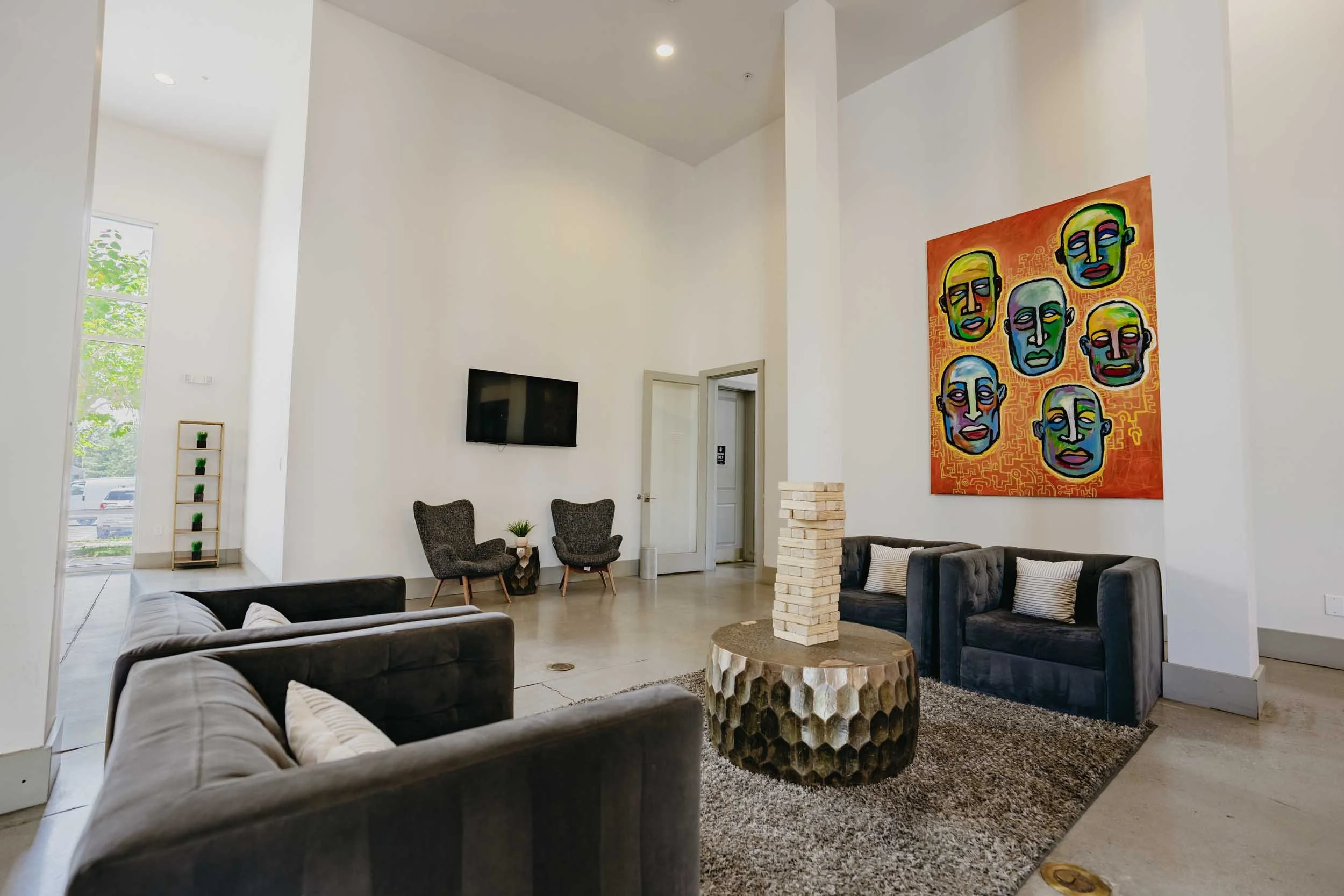 A modern, minimalist waiting area with dark gray sofas, a wooden coffee table with a unique honeycomb design, two armchairs, a large colorful abstract portrait painting of multiple faces, a flat-screen TV, and a small shelf with potted plants, in a b