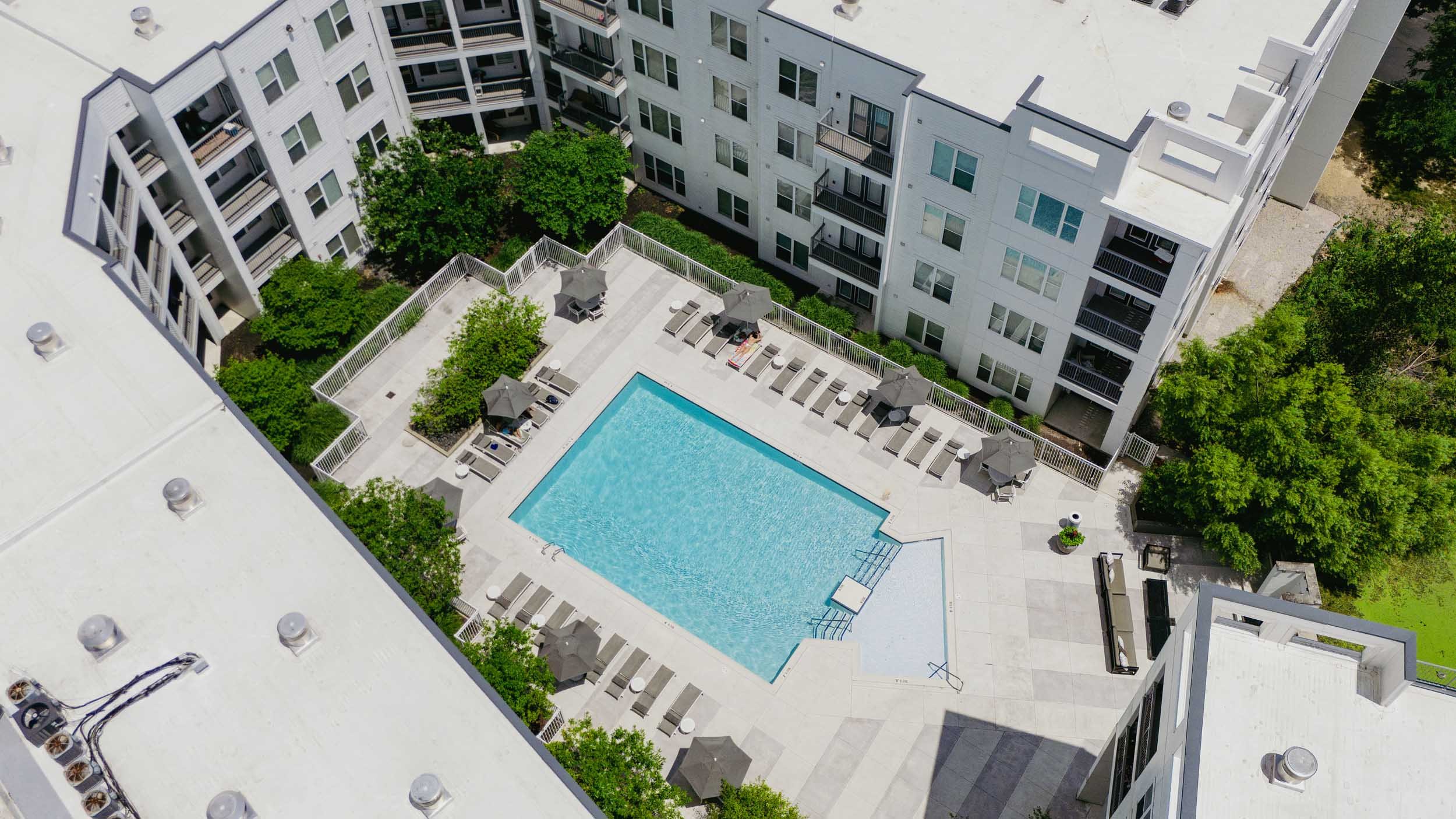 A view from above of a residential apartment complex with a swimming pool, surrounded by lounge chairs, umbrellas, and green trees.