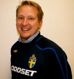 A man smiling, wearing a navy blue sports jersey with a Swedish flag emblem and sponsor logo, standing against a plain wall.