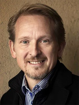 A close-up portrait of a man with light skin, medium-length light brown hair, a goatee, and a mustache, smiling and wearing a dark jacket over a striped shirt, against a plain background.