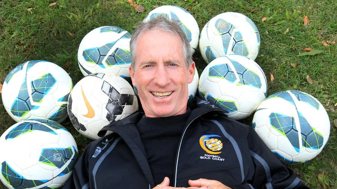 Man lying on grass surrounded by multiple yellow and white soccer balls, wearing a black jacket with a 'Football Gold Coast' logo.