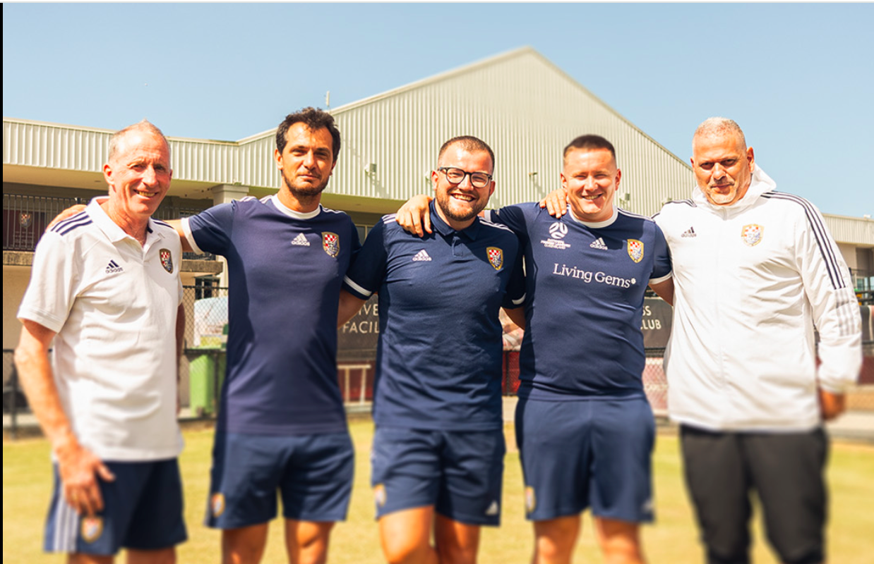 Five men posing together outdoors, some wearing matching navy blue sports jerseys, with two in white jackets, standing in front of a sports facility, smiling under a clear blue sky.