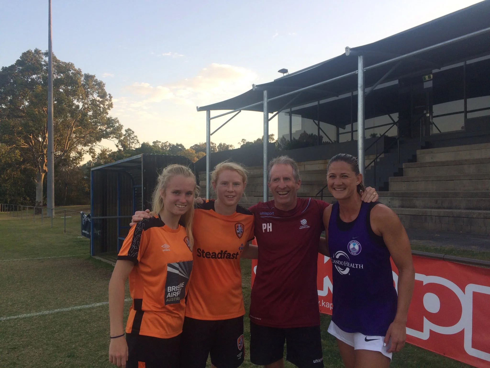 Four people, two young women and two adults, standing on a sports field after a game, smiling and with their arms around each other, with trees and a stadium seating area in the background.