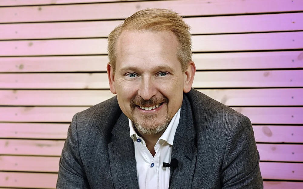 Smiling man with blond hair and beard wearing a gray suit jacket and white shirt, sitting in front of a pink wooden slat wall.